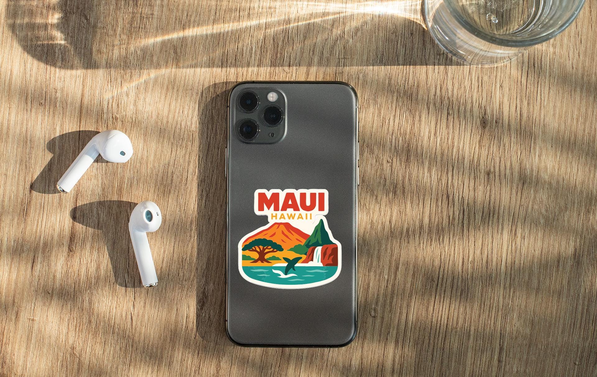 a smartphone with a sticker of a Hawaiian landscape, a pair of white earbuds, and a glass of water on a wooden surface.