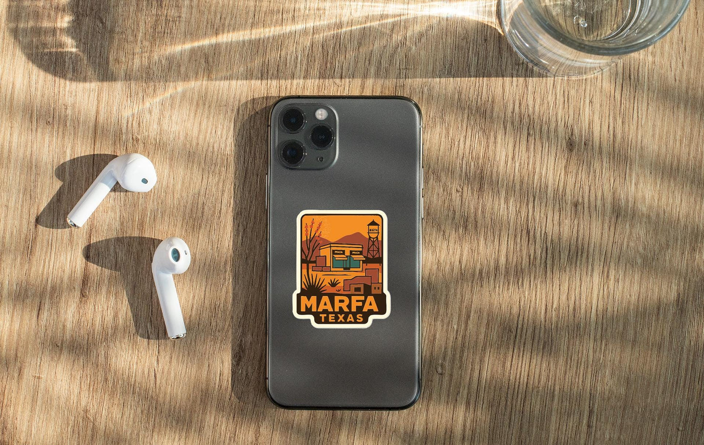 a smartphone with a sticker on the back, placed on a wooden surface alongside a pair of white earbuds and a glass of water.