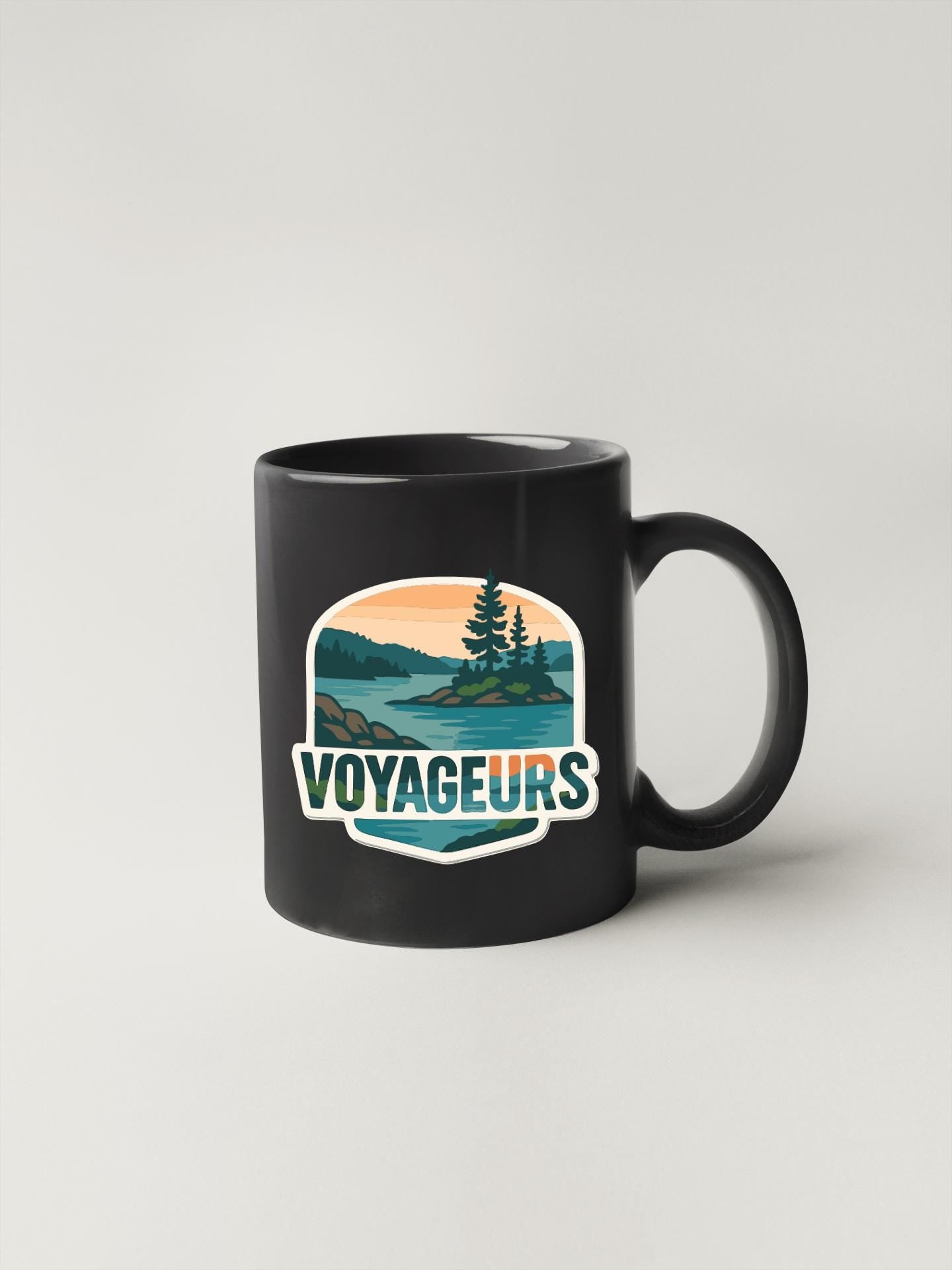 A black coffee mug with a graphic design depicting a scenic landscape with a lake, mountains, and trees. The text "VOYAGEURS" is displayed prominently on the mug.
