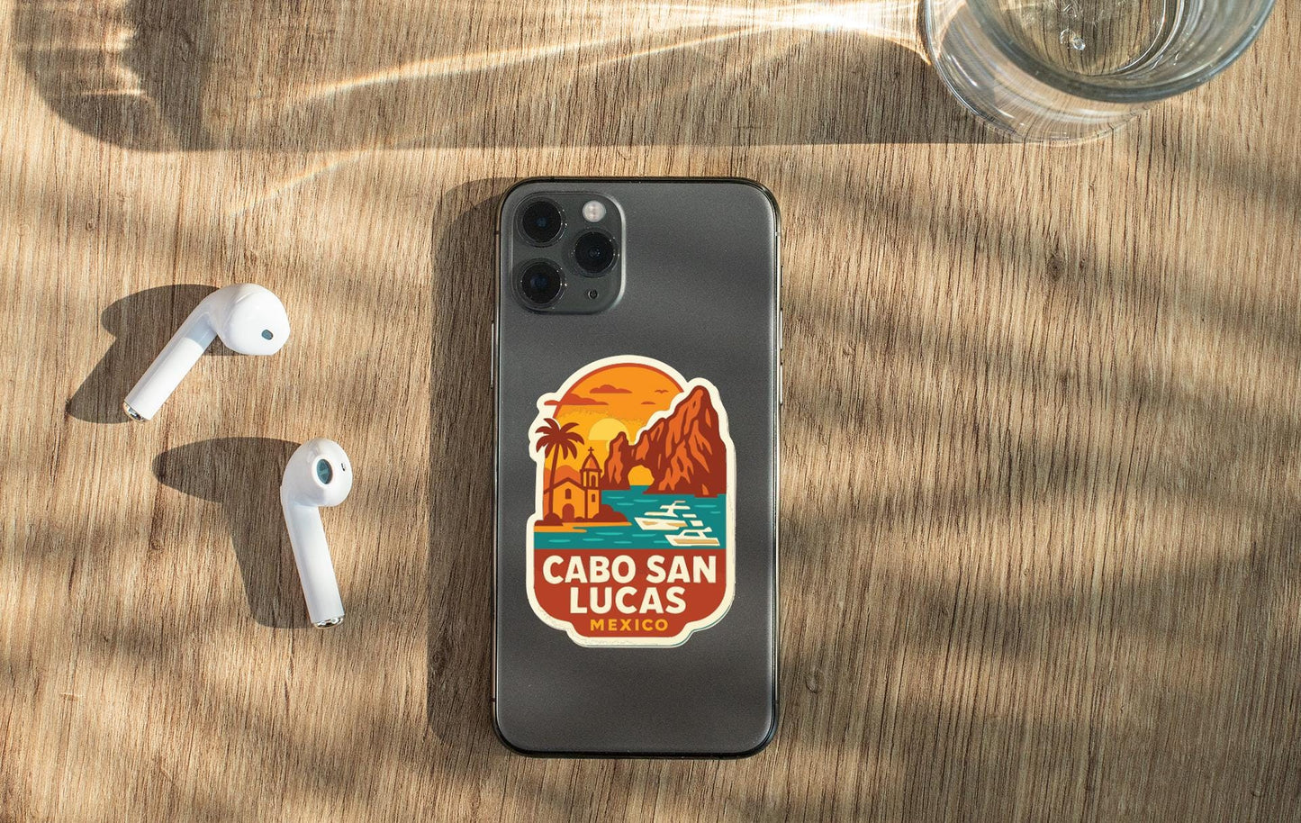 a smartphone with a sticker of Cabo San Lucas, Mexico, placed on a wooden surface alongside a pair of white earbuds and a glass of water.