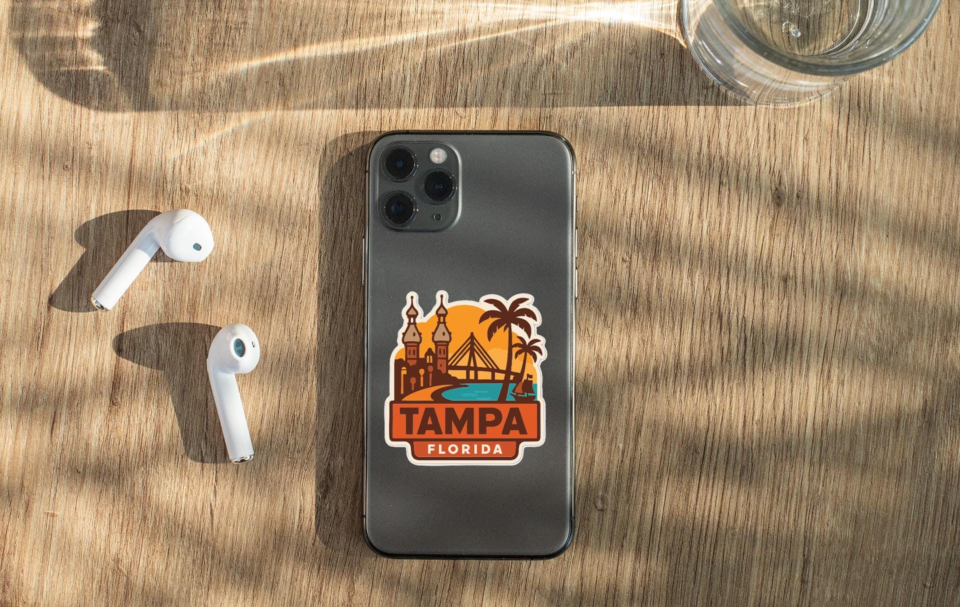 a smartphone with a sticker of Tampa, Florida, placed on a wooden surface alongside a pair of white earbuds and a glass of water.