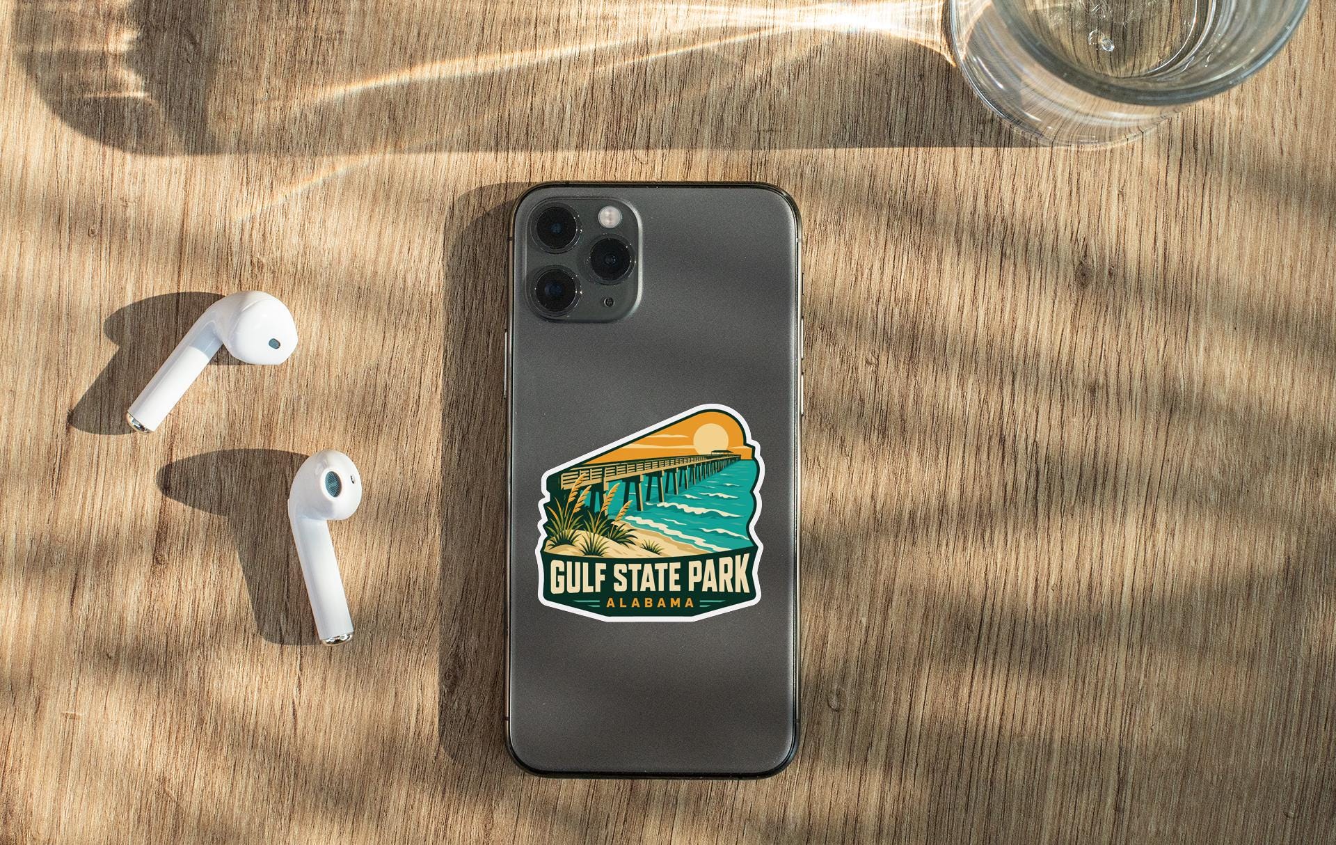 a smartphone with a sticker on it, placed on a wooden surface alongside a pair of white earbuds and a glass of water.