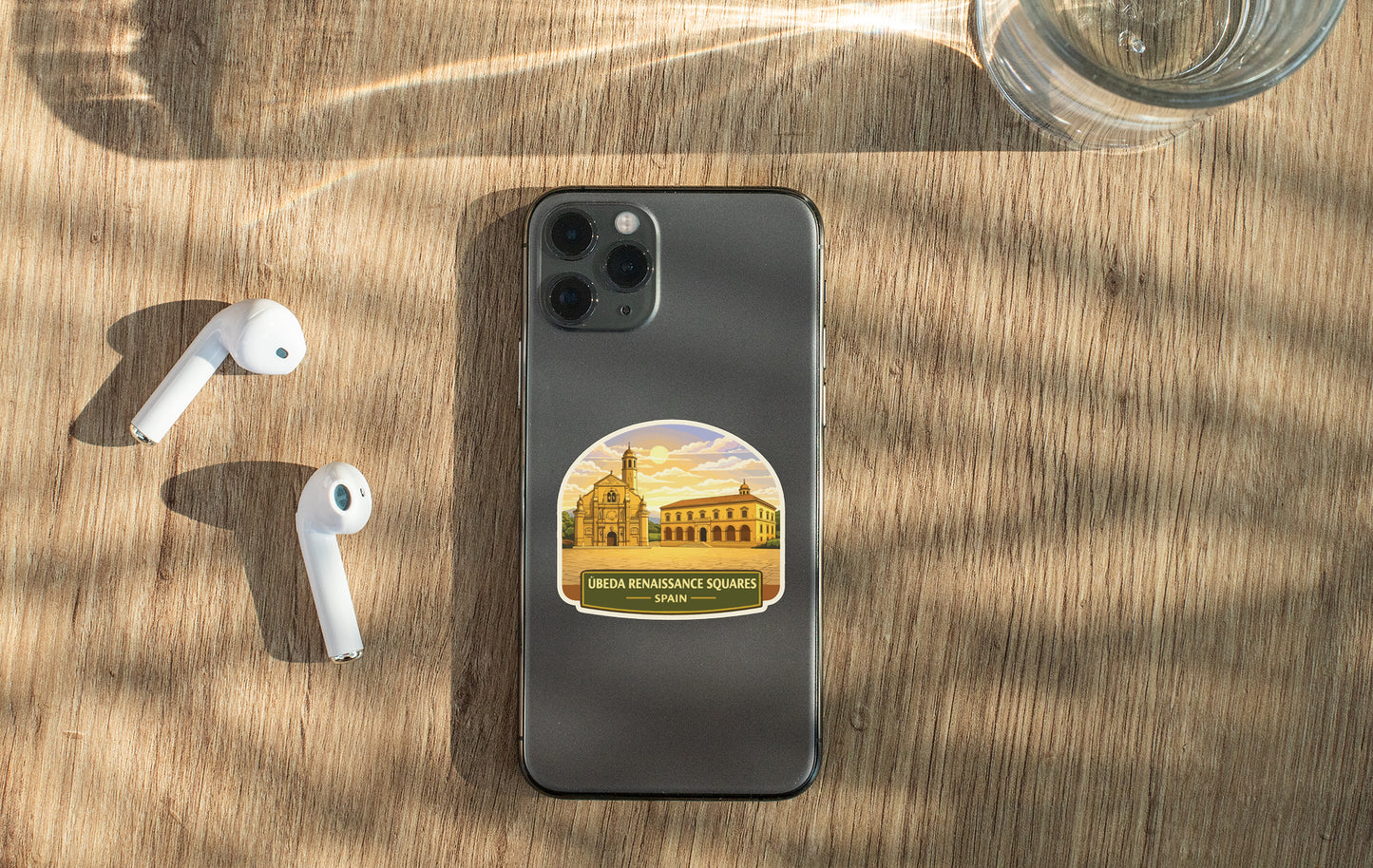 a smartphone with a sticker of a building on it, placed on a wooden surface alongside a pair of white earbuds and a glass of water.