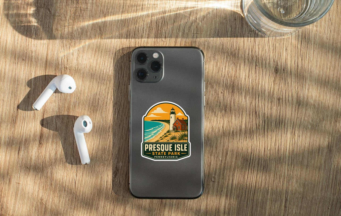 a smartphone with a sticker on it, placed on a wooden surface alongside a pair of white earbuds and a glass of water.