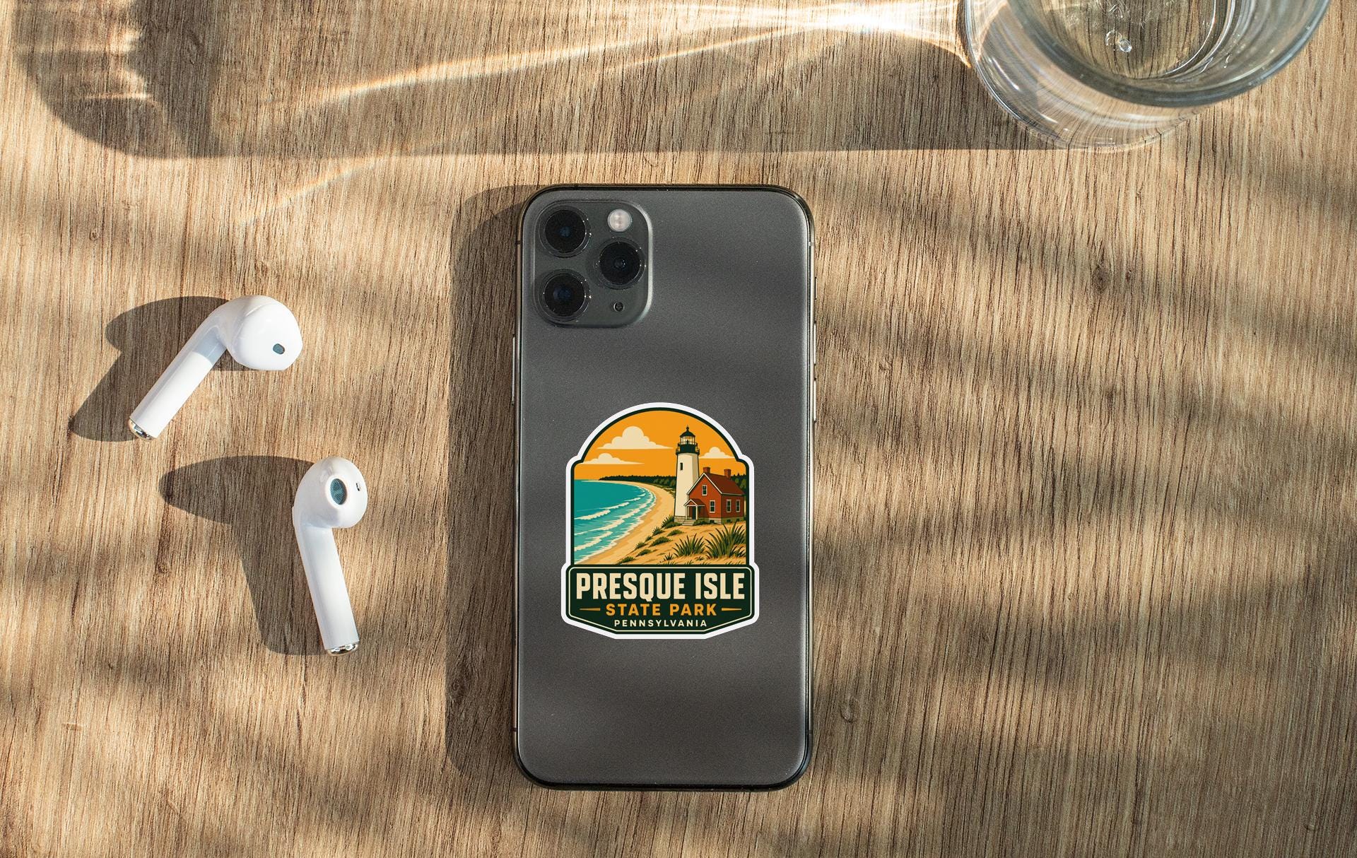 a smartphone with a sticker on it, placed on a wooden surface alongside a pair of white earbuds and a glass of water.