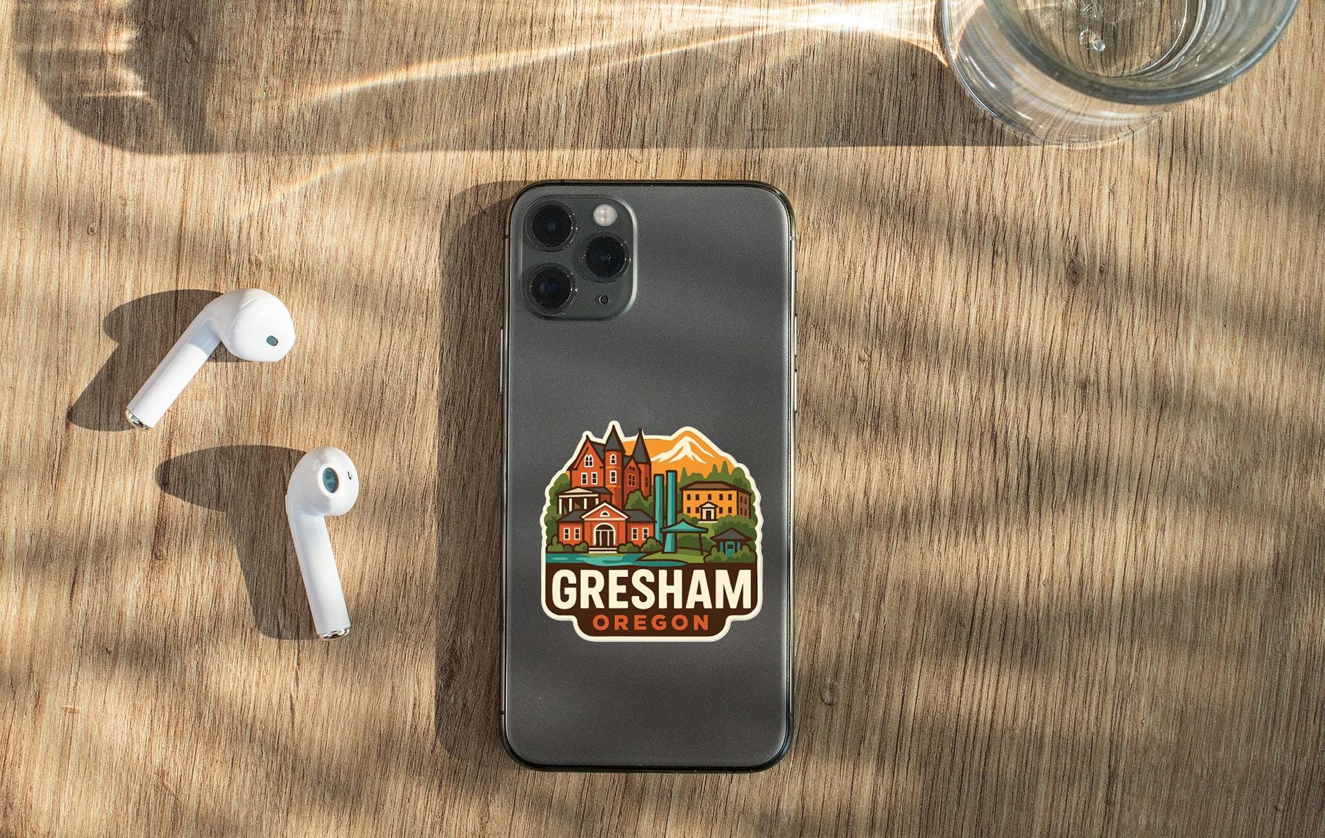 a smartphone with a sticker on the back, placed on a wooden surface alongside a pair of white earbuds and a glass of water.