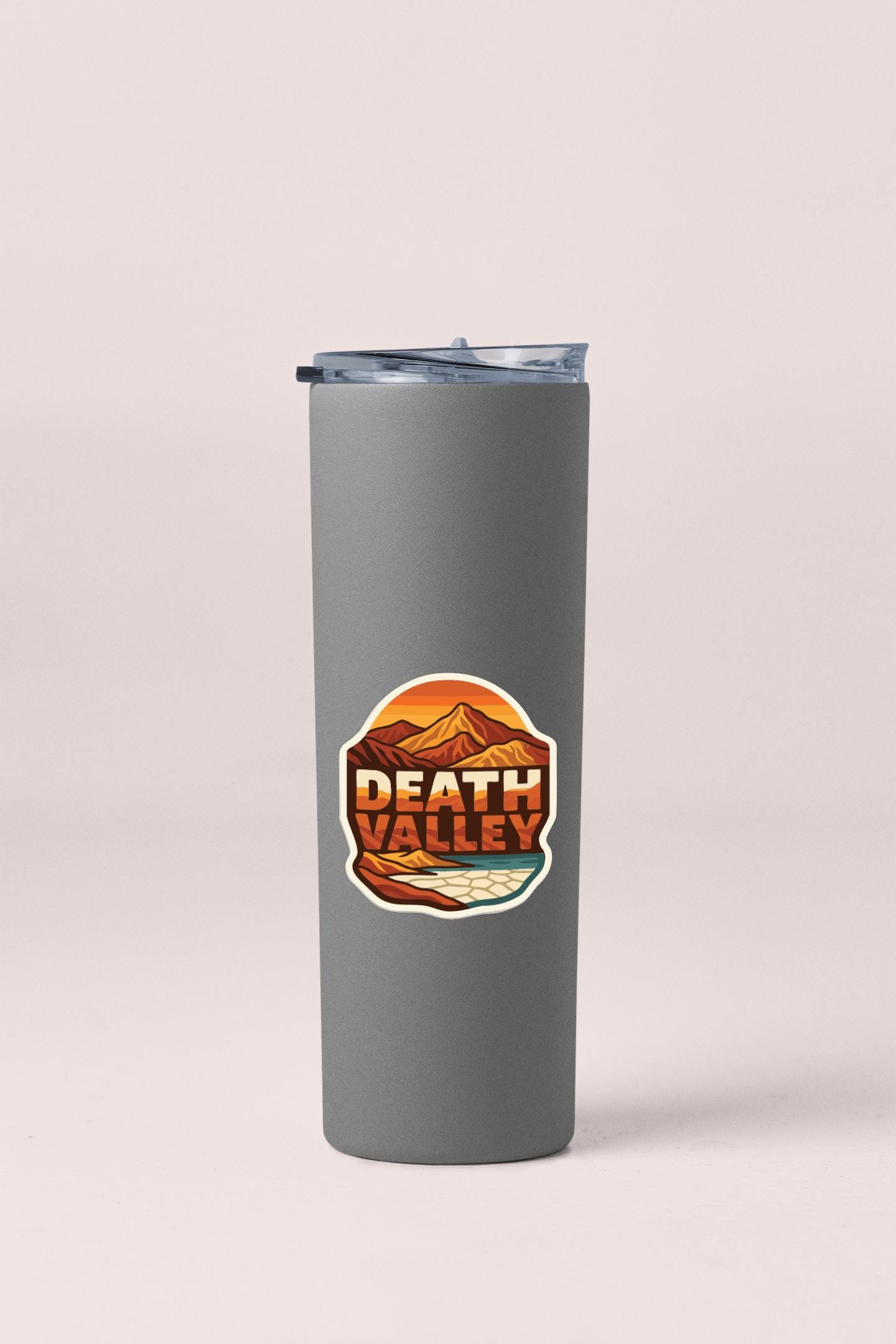 A gray insulated tumbler with a lid and a circular sticker featuring an illustration of mountains and a body of water.