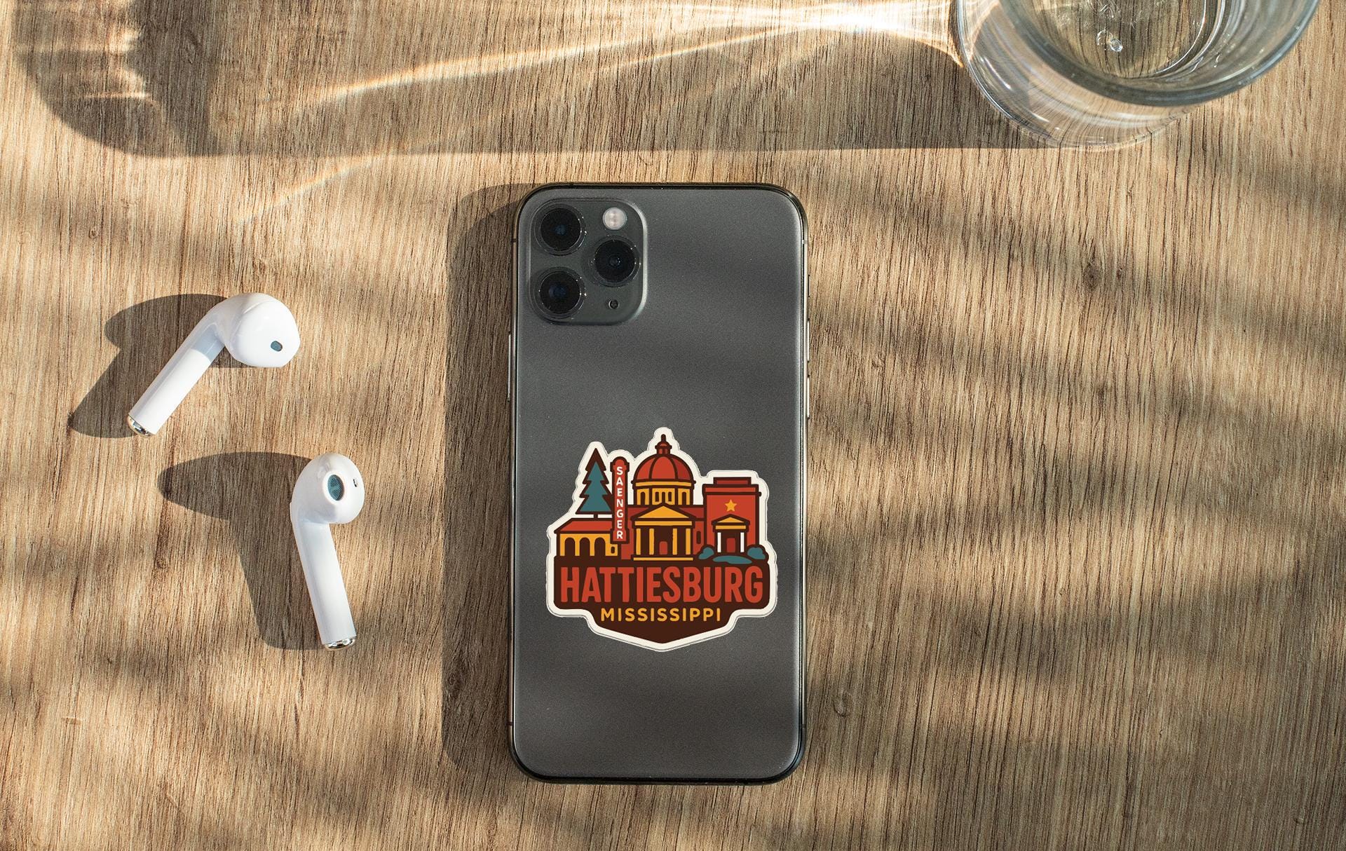 a smartphone with a sticker of Hattiesburg, Mississippi, placed on a wooden surface alongside a pair of white earbuds and a glass of water.