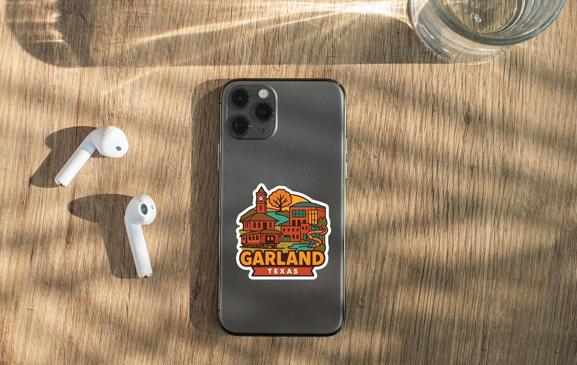 a smartphone with a sticker of the state of Texas on its back, along with a pair of white earbuds and a glass of water on a wooden surface.