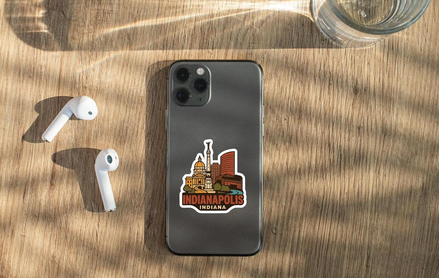 a smartphone with a sticker of Indianapolis, Indiana, placed on a wooden surface alongside a pair of white earbuds and a glass of water.