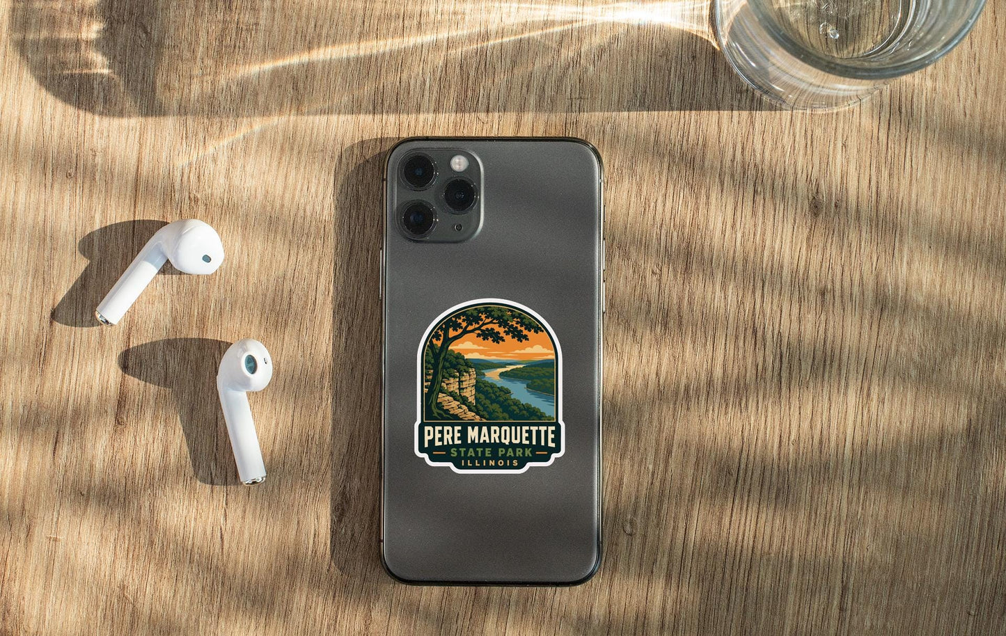 a smartphone with a sticker on the back, placed on a wooden surface alongside a pair of white earbuds and a glass of water.
