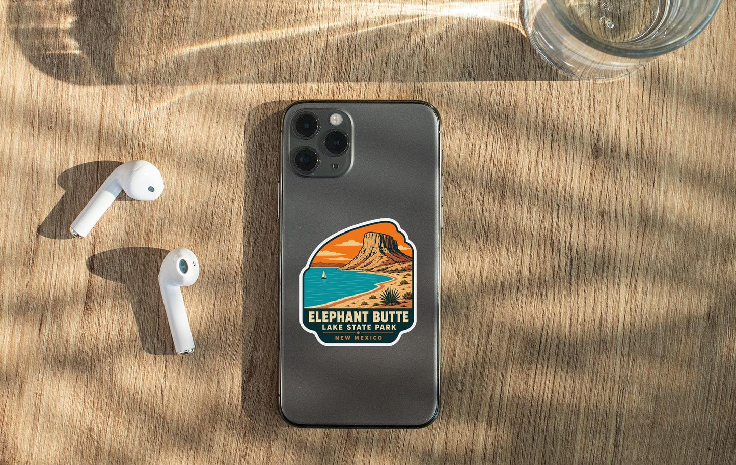 a smartphone with a sticker on the back, placed on a wooden surface alongside a pair of white earbuds and a glass of water.