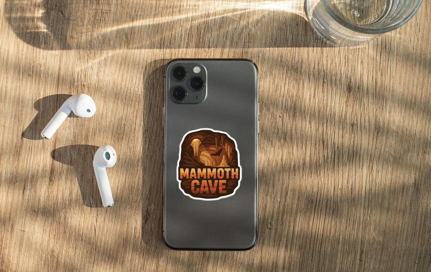 a smartphone with a sticker on it, placed on a wooden surface alongside a pair of white earbuds and a glass of water.