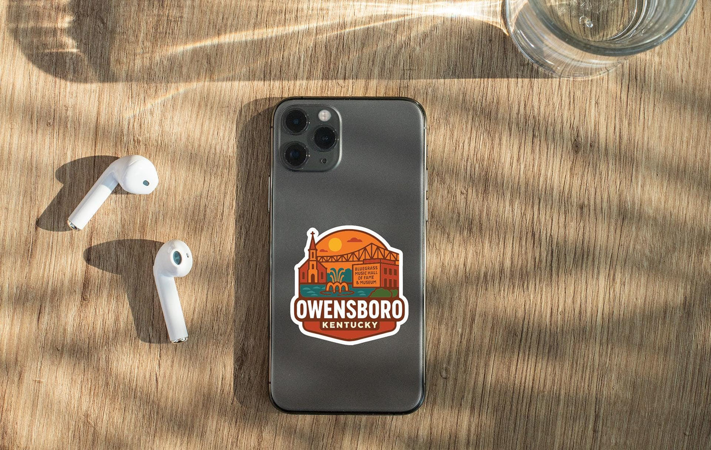 a smartphone with a sticker on it, placed on a wooden surface alongside a pair of white earbuds and a glass of water.