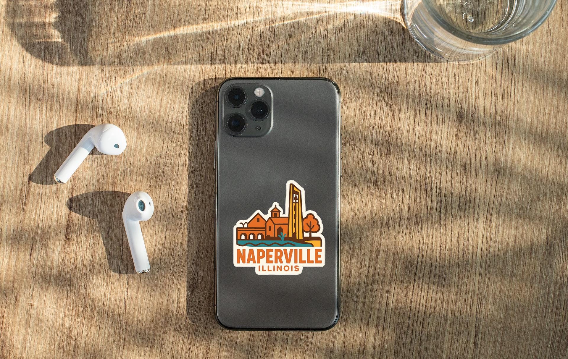a smartphone with a sticker of Naperville Illinois on it, along with a pair of white earbuds and a glass of water on a wooden surface.