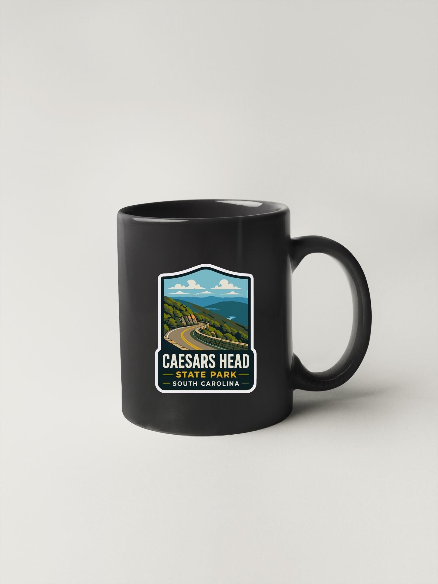 A black coffee mug with a sticker depicting a scenic landscape, including a winding road and mountains in the background.