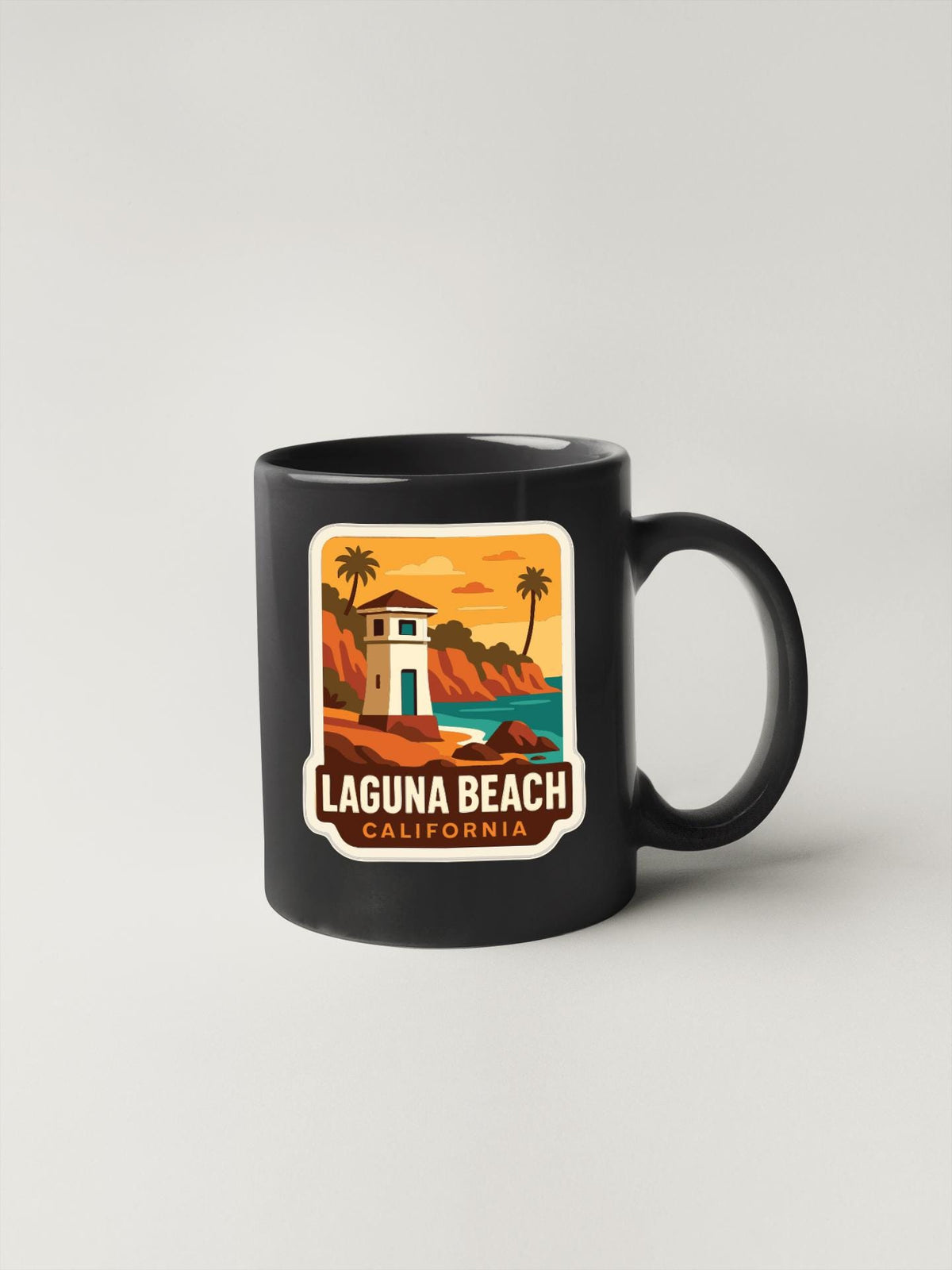 A black coffee mug with a sticker depicting a beach scene and the text "LAGUNA BEACH CALIFORNIA".