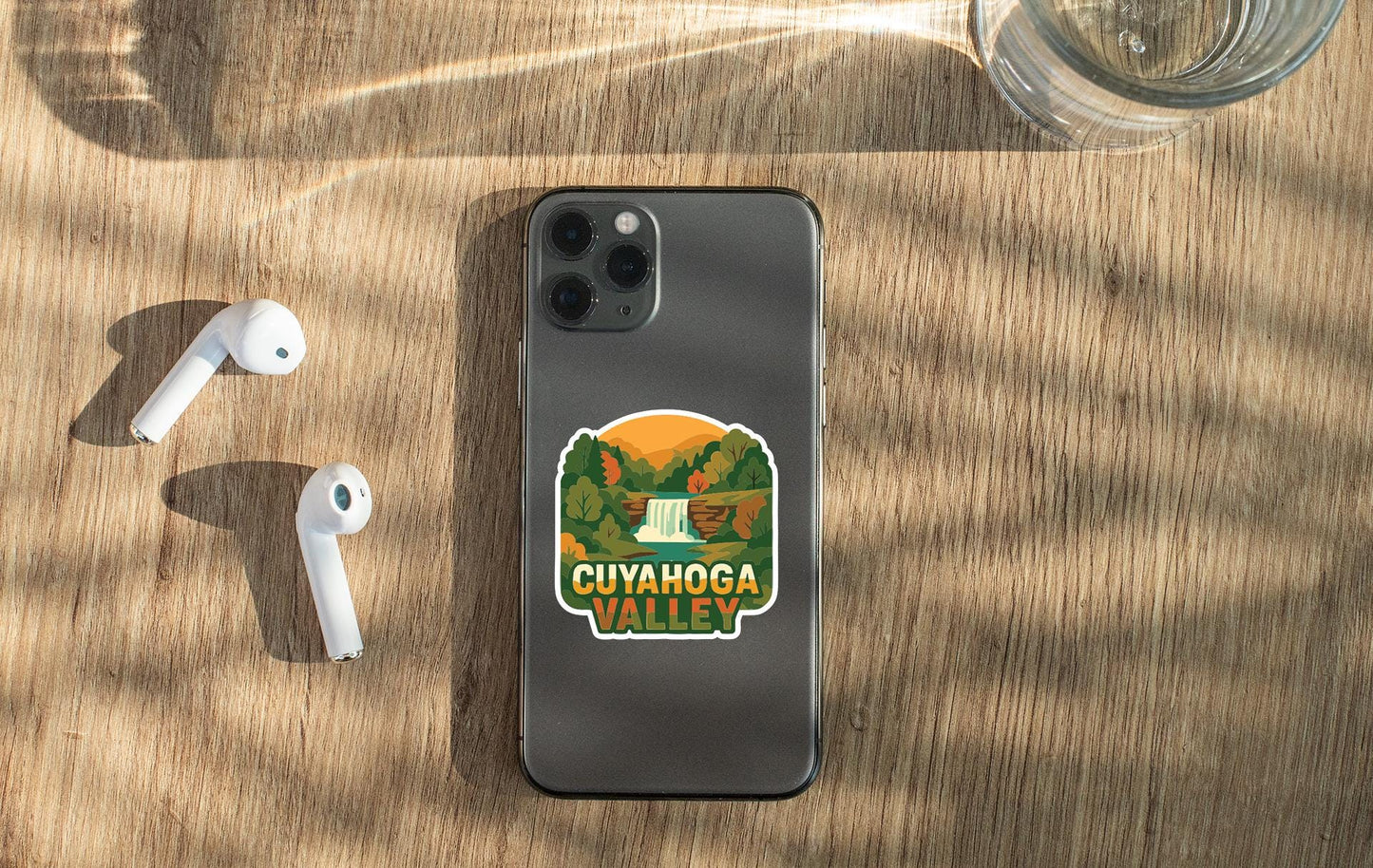 a smartphone with a sticker on the back, along with a pair of white earbuds and a glass of water on a wooden surface.