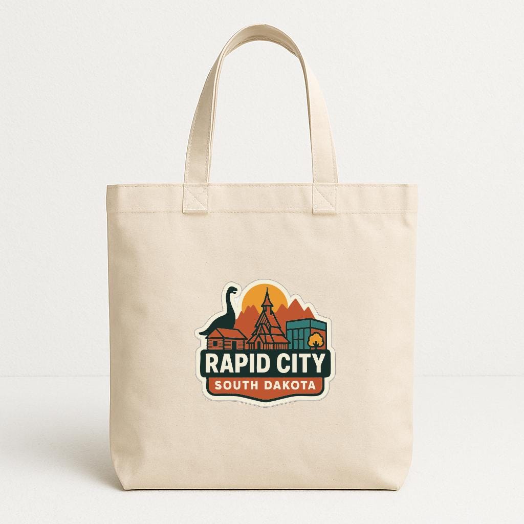 A beige tote bag with a graphic design featuring a dinosaur, buildings, and mountains, along with the text "RAPID CITY SOUTH DAKOTA".