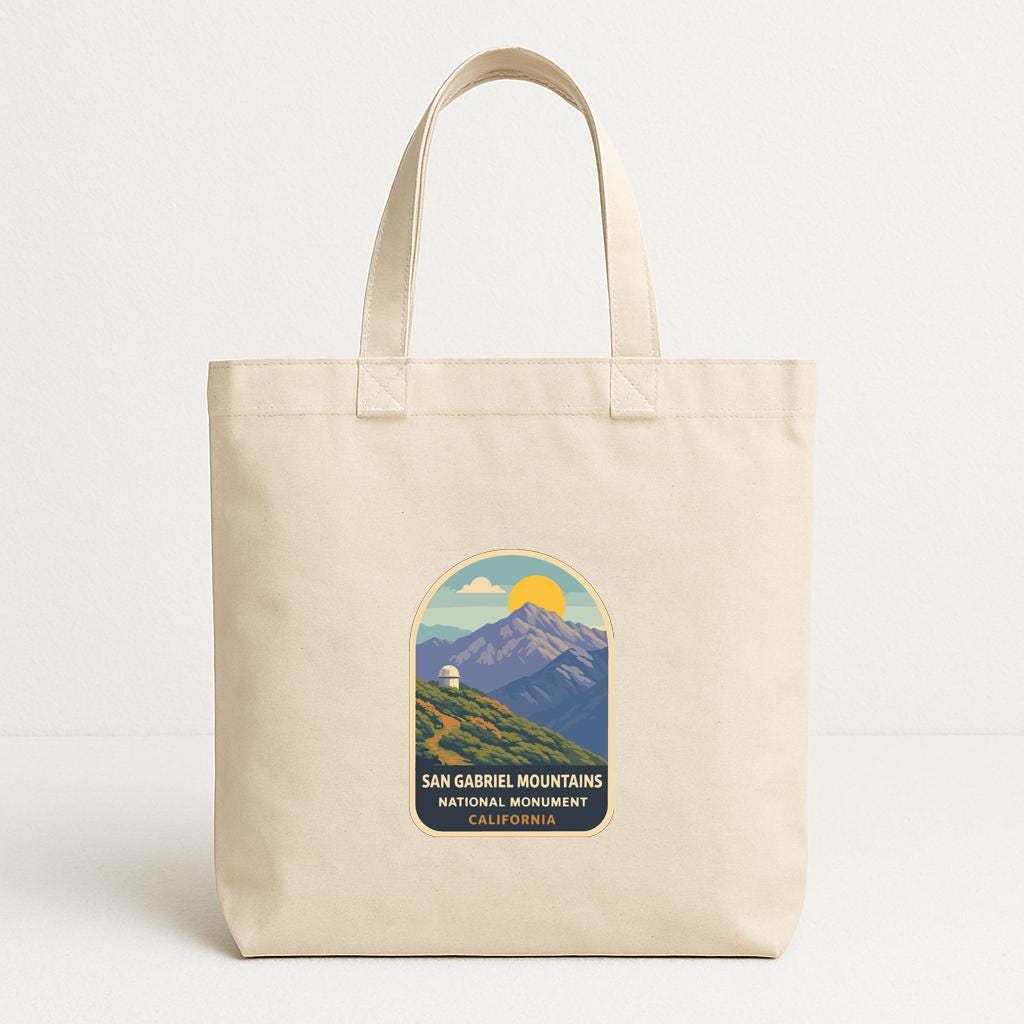 A beige tote bag with a graphic design featuring a mountain landscape and the text "SAN GABRIEL MOUNTAINS NATIONAL MONUMENT CALIFORNIA" printed on it.