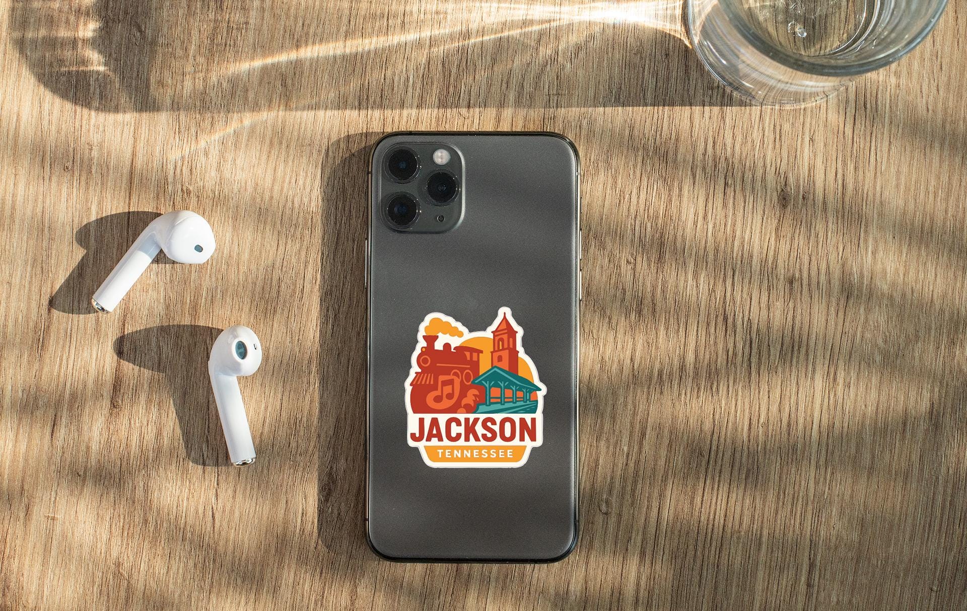 a smartphone with a sticker on it, placed on a wooden surface alongside a pair of white earbuds and a glass of water.