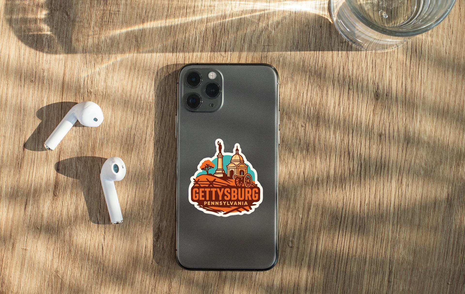 a smartphone with a sticker of Gettysburg, Pennsylvania, on its back, along with a pair of white earbuds and a glass of water on a wooden surface.