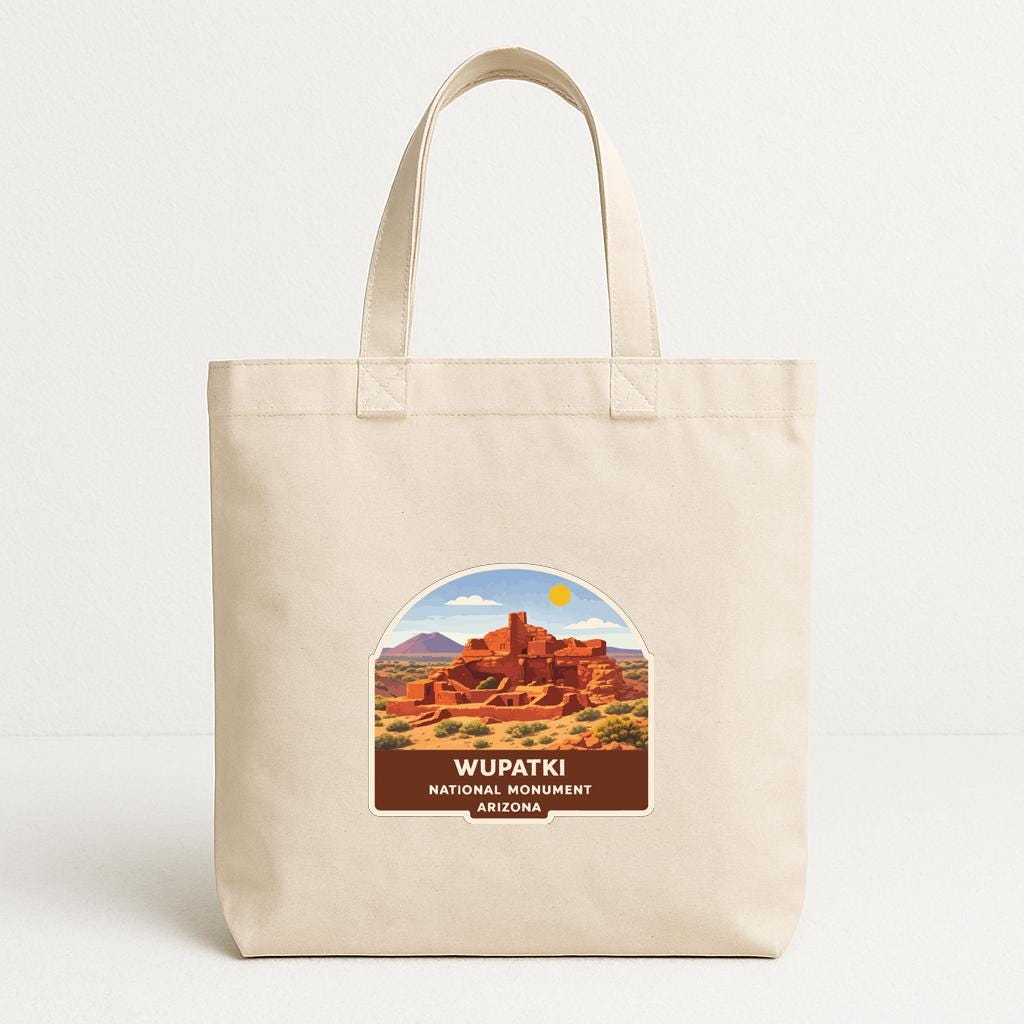 A beige tote bag with a printed label that says "WUPATKI NATIONAL MOUNTAIN ARIZONA" and features an illustration of a desert landscape with a red rock formation.