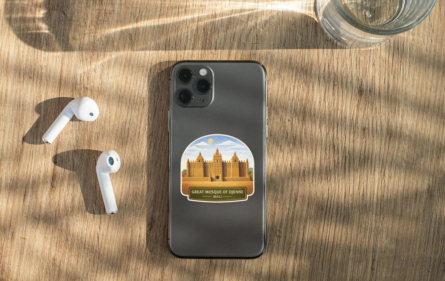 a smartphone with a sticker of the Great Mosque of Djenné, a UNESCO World Heritage Site in Mali, placed on a wooden surface alongside a pair of white earbuds and a glass of water.