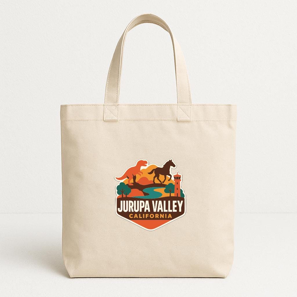 A beige tote bag with a graphic design featuring a dinosaur, horse, and mountains, along with the text "JURUPA VALLEY CALIFORNIA".