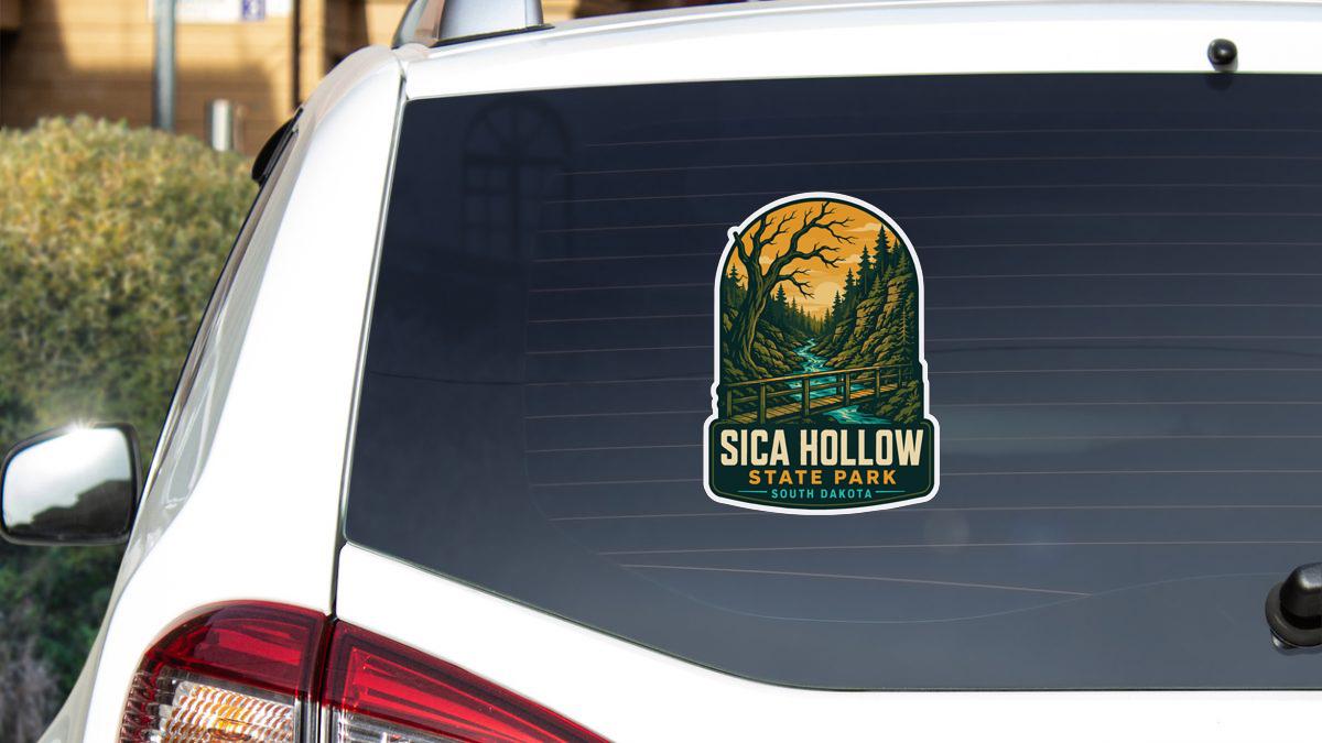 A sticker on the rear window of a white car, depicting a scenic landscape with a bridge and trees, advertising the location of Sica Hollow State Park in South Dakota.