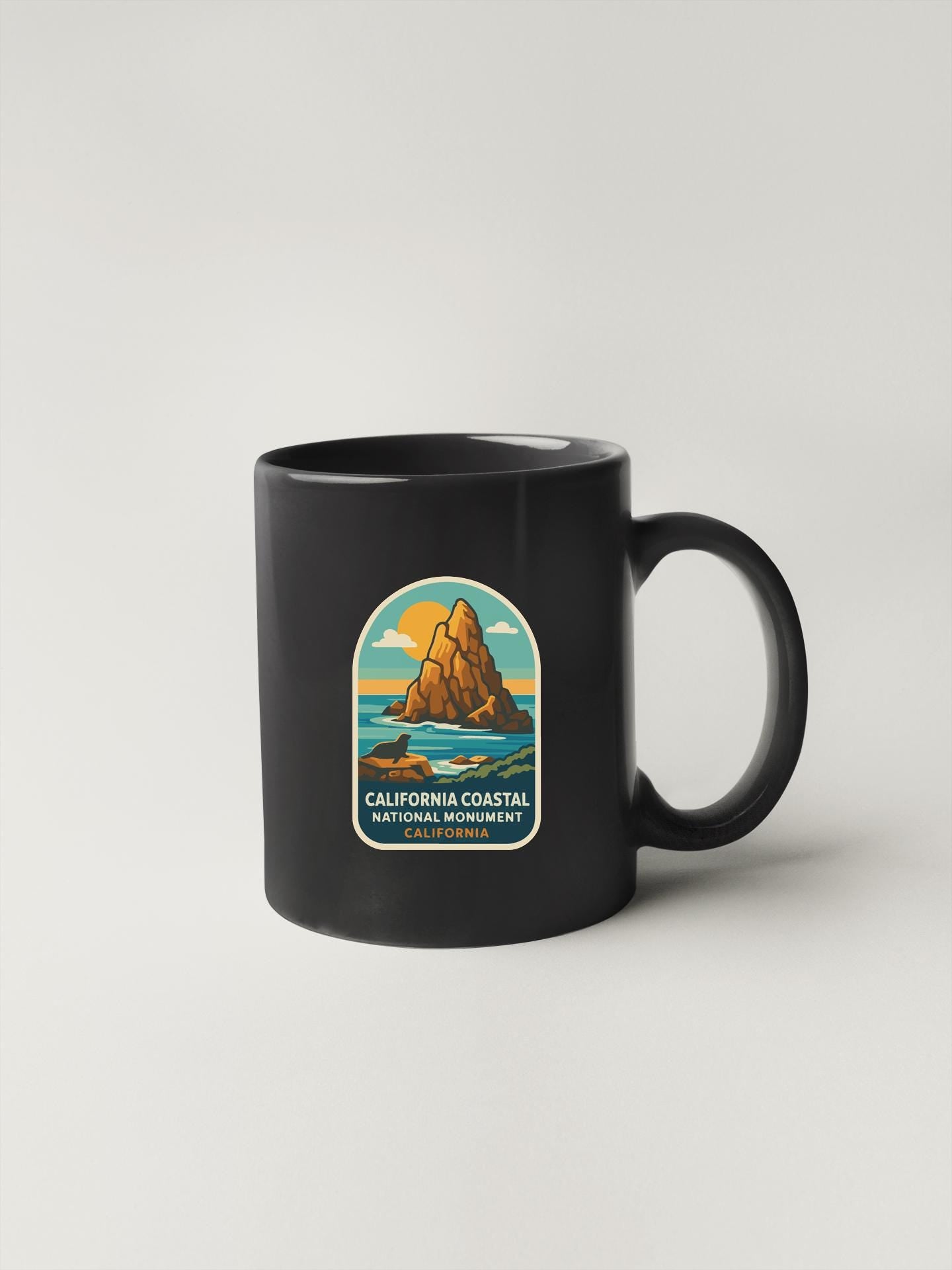 A black coffee mug with a graphic design depicting a coastal scene, including a mountain, ocean, and sunset.