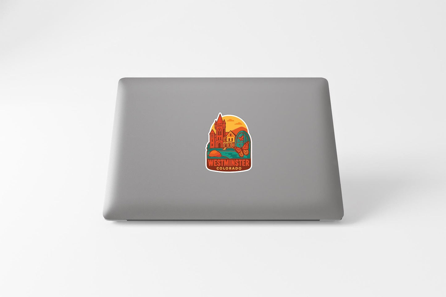 A sticker depicting a colorful illustration of a landmark, likely Westminster, Colorado, is placed on the back of a laptop computer.