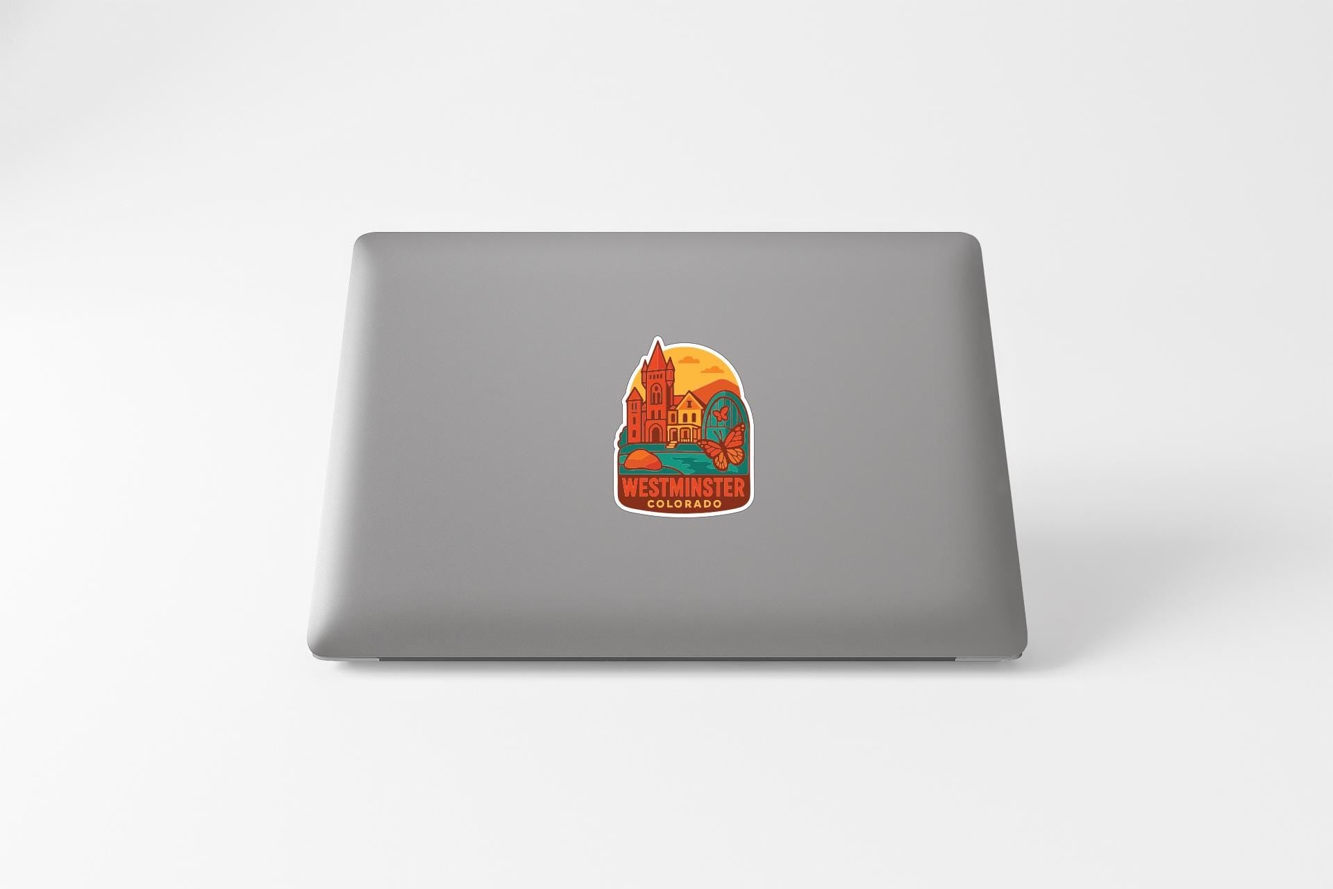 A sticker depicting a colorful illustration of a landmark, likely Westminster, Colorado, is placed on the back of a laptop computer.