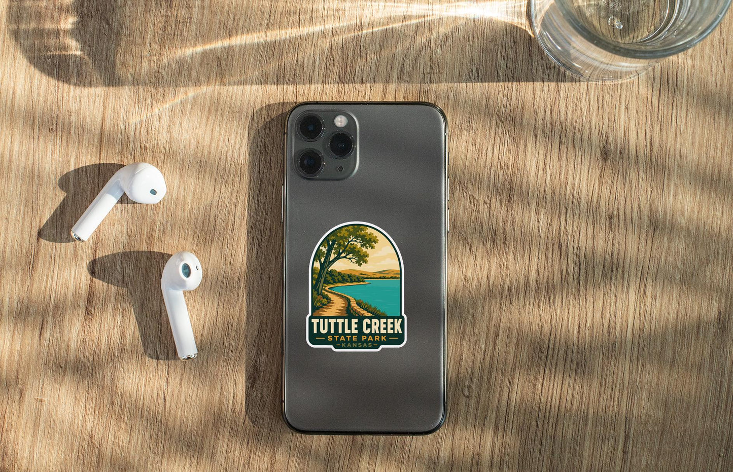 a smartphone with a sticker on the back, placed on a wooden surface alongside a pair of white earbuds and a glass of water.