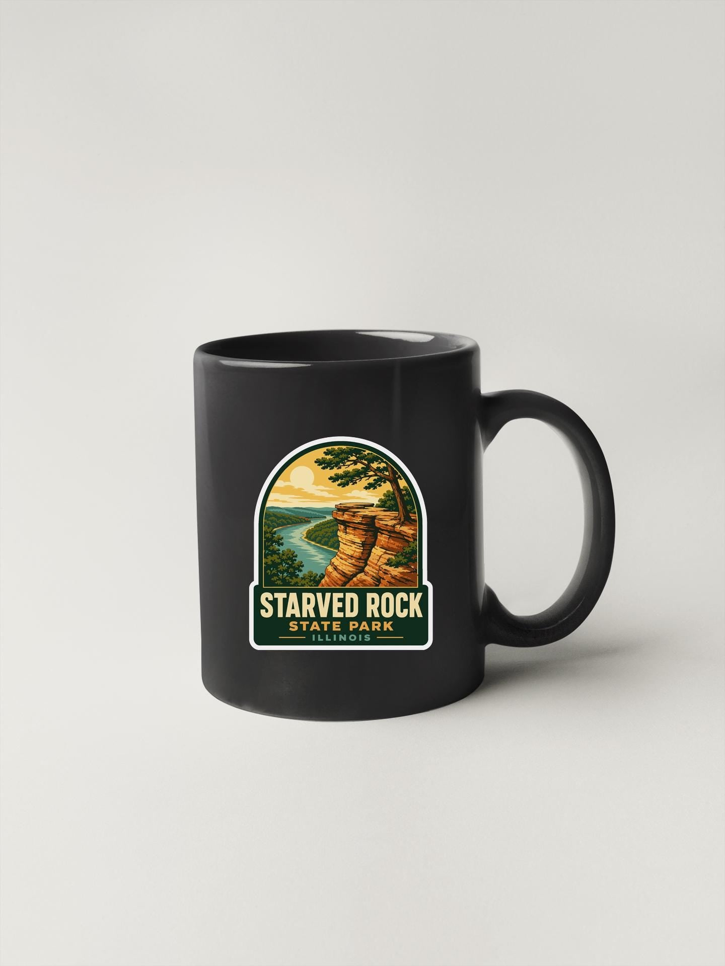 A black coffee mug with a sticker depicting a scenic landscape featuring a cliff overlooking a river.