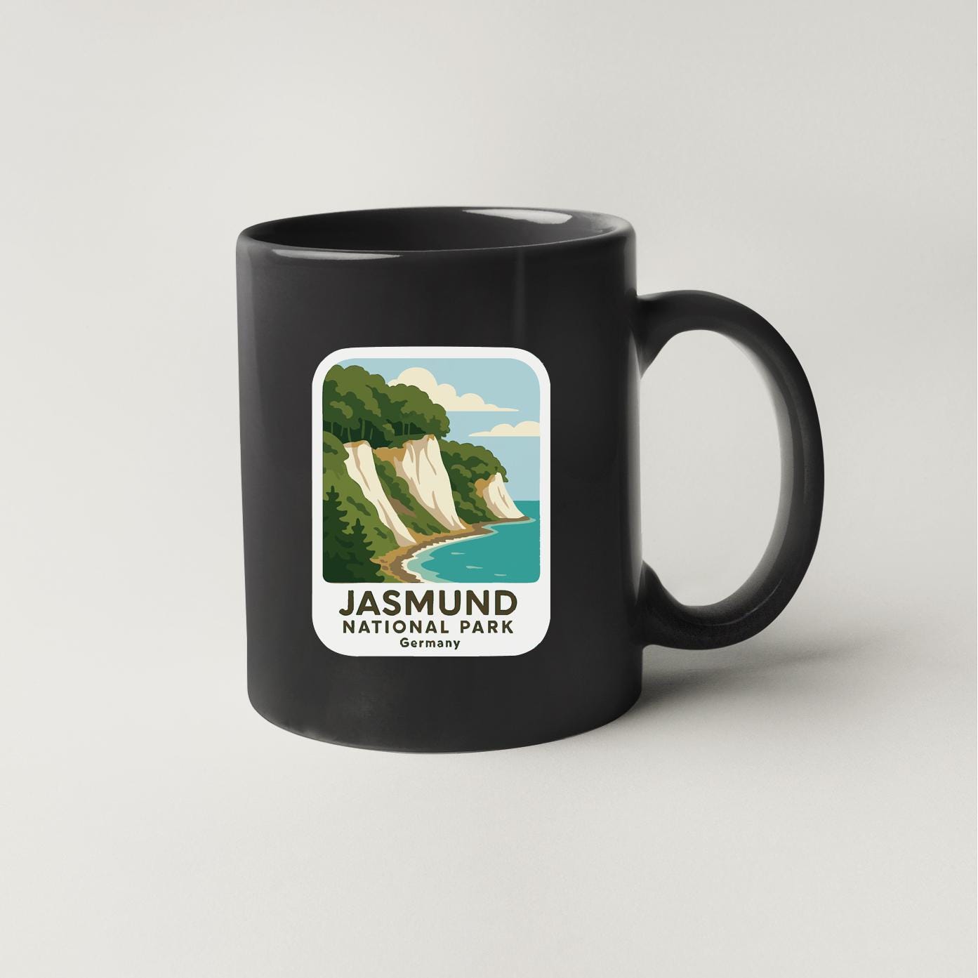 A black coffee mug with a graphic design depicting a scenic coastal landscape, including cliffs, a beach, and a body of water.