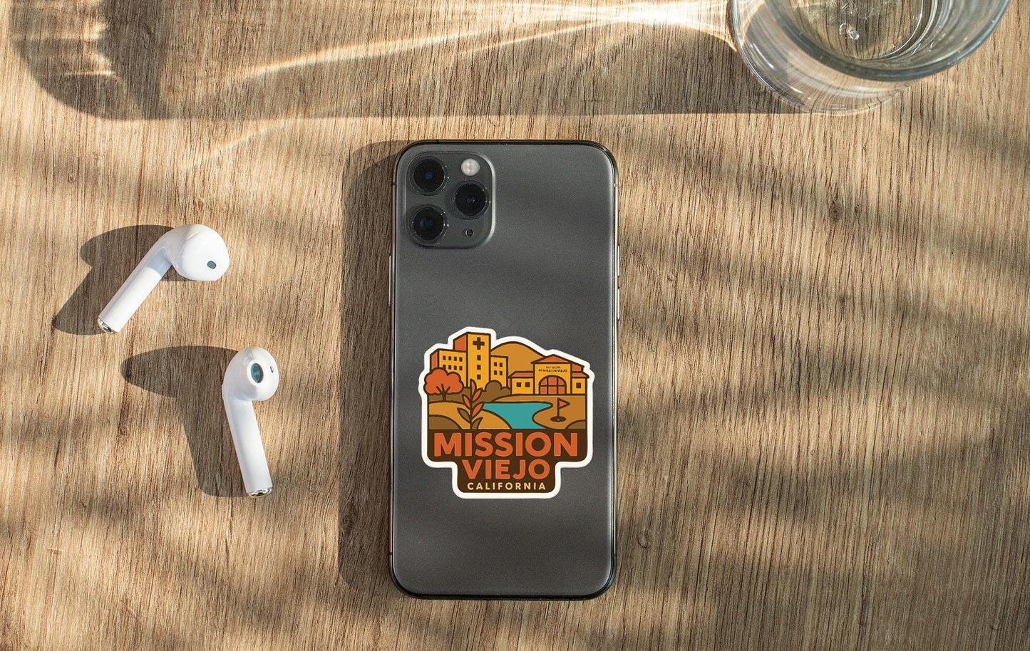 a smartphone with a sticker on the back, placed on a wooden surface alongside a pair of white earbuds and a glass of water.