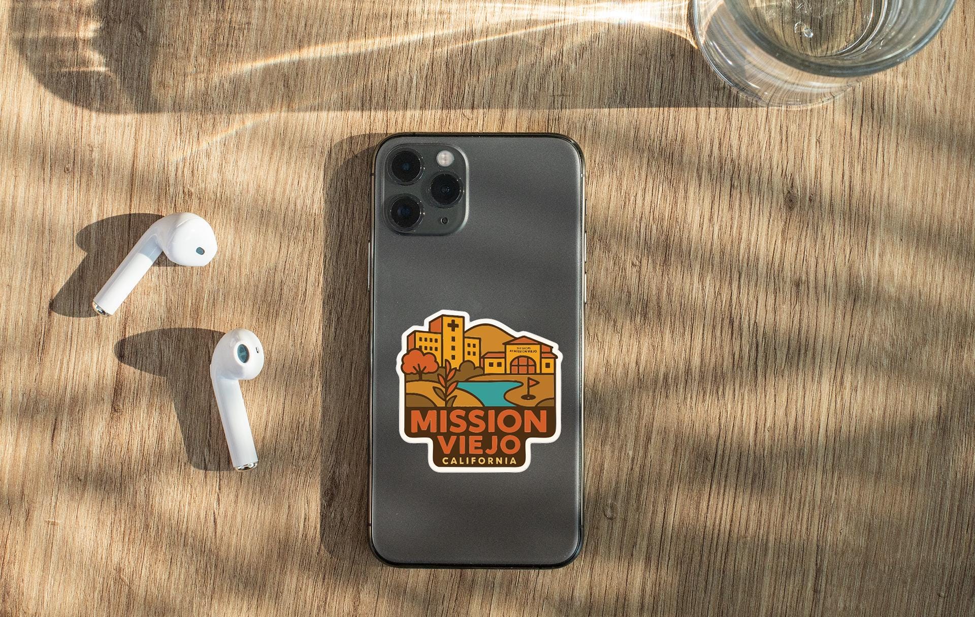 a smartphone with a sticker on the back, placed on a wooden surface alongside a pair of white earbuds and a glass of water.
