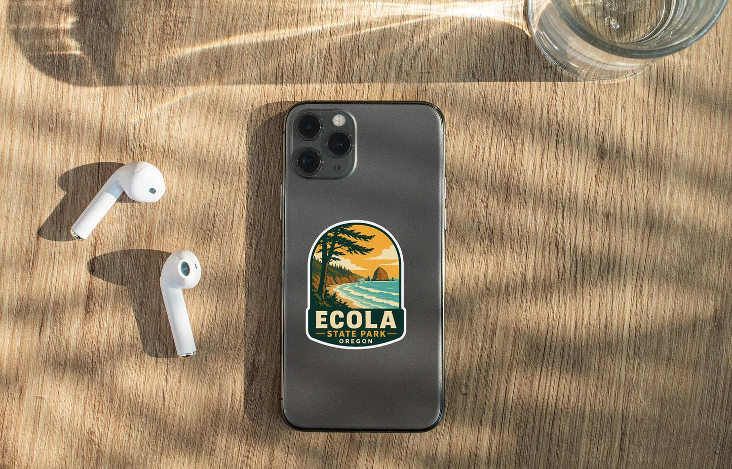 a smartphone with a sticker on it, placed on a wooden surface alongside a pair of white earbuds and a glass of water.