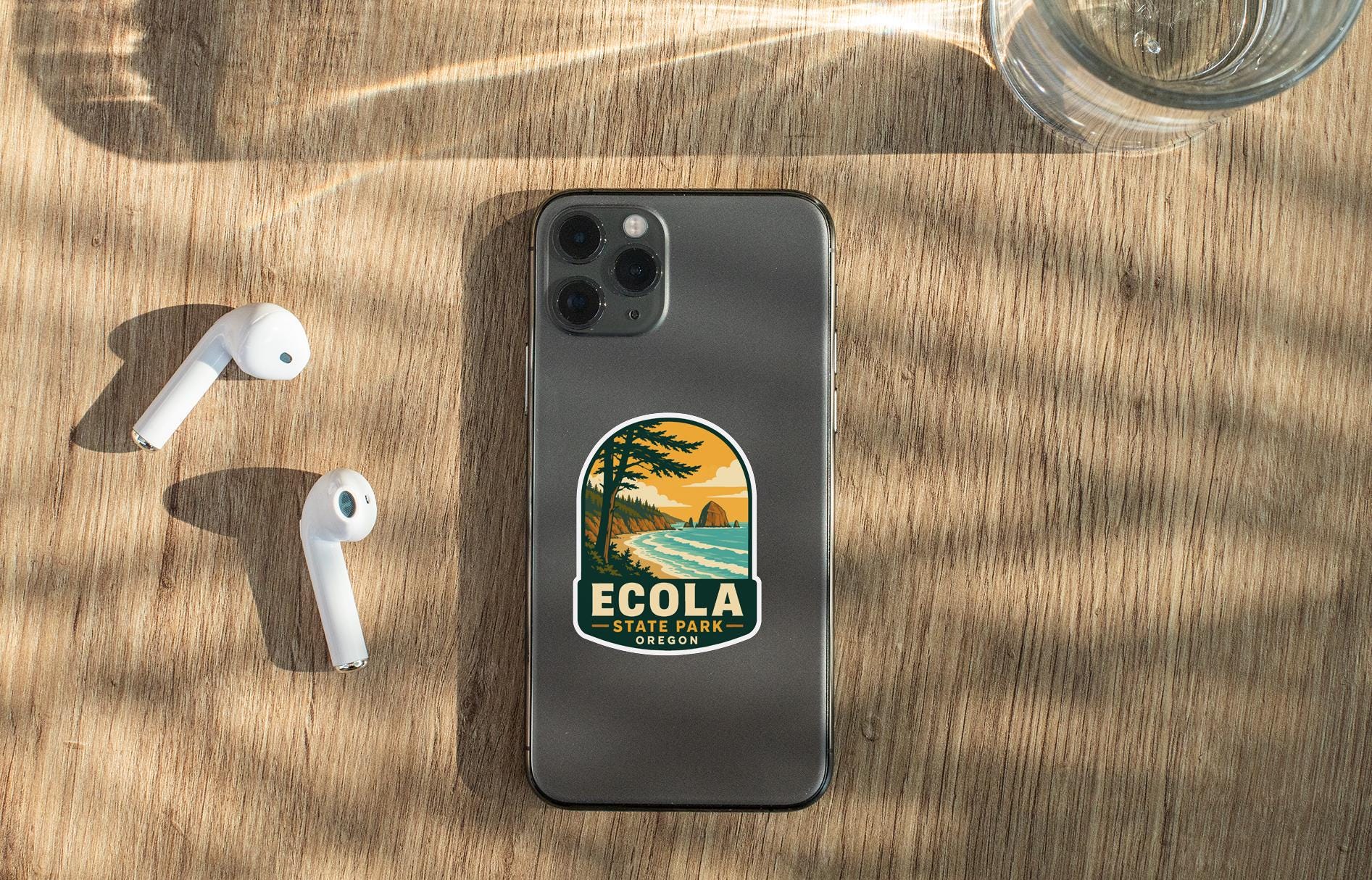a smartphone with a sticker on it, placed on a wooden surface alongside a pair of white earbuds and a glass of water.