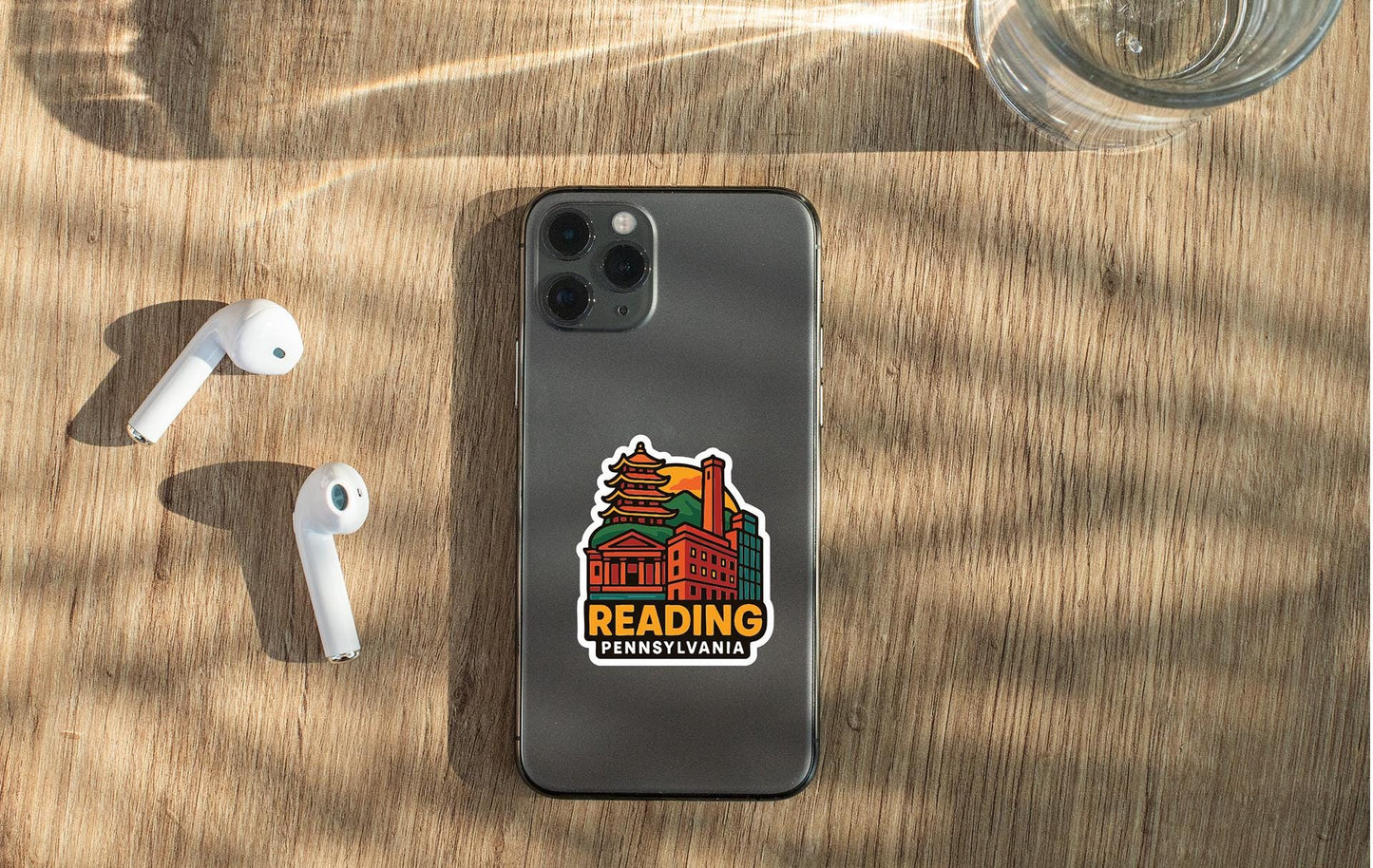 a smartphone with a sticker on the back, placed on a wooden surface alongside a pair of white earbuds and a glass of water.
