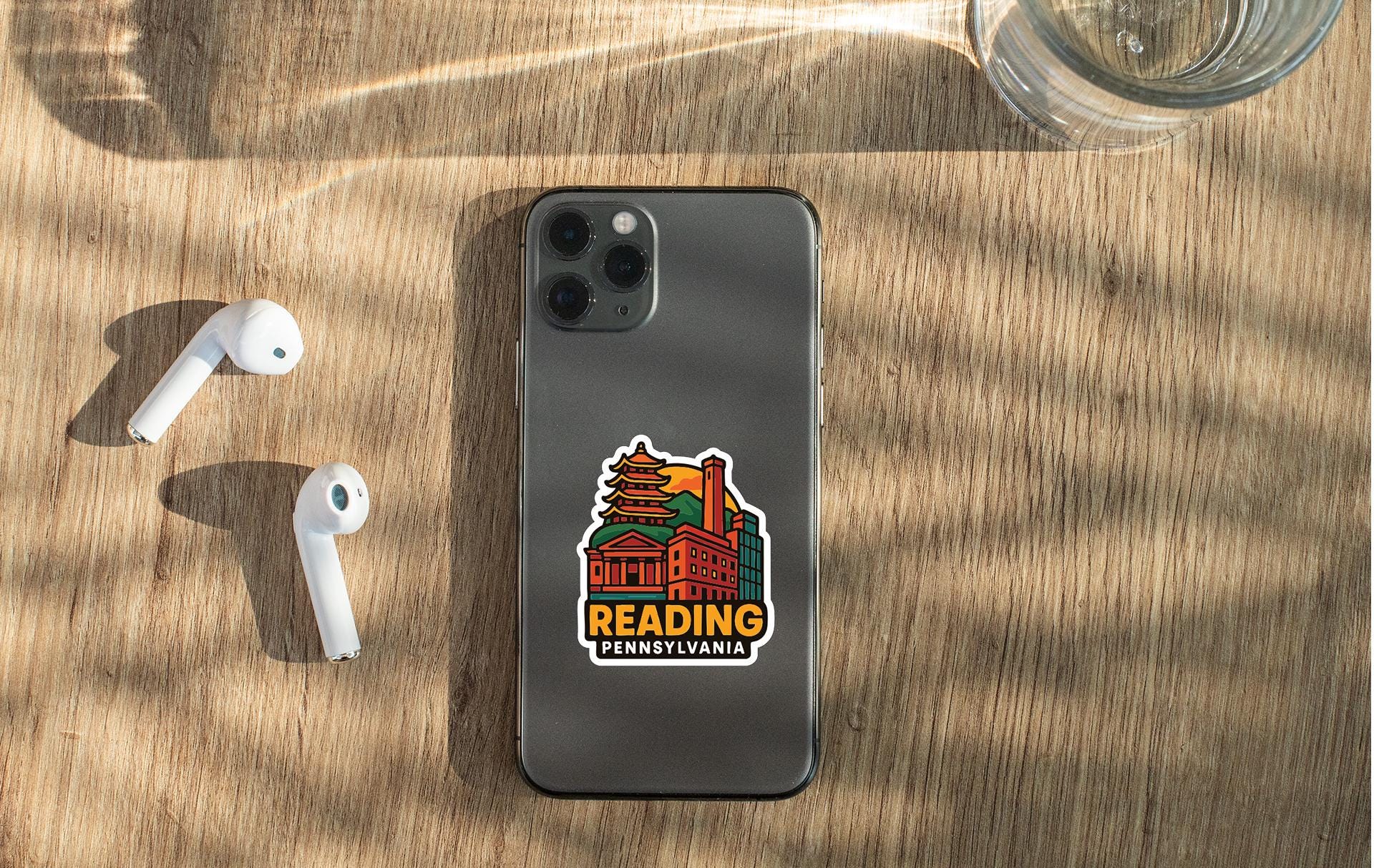 a smartphone with a sticker on the back, placed on a wooden surface alongside a pair of white earbuds and a glass of water.