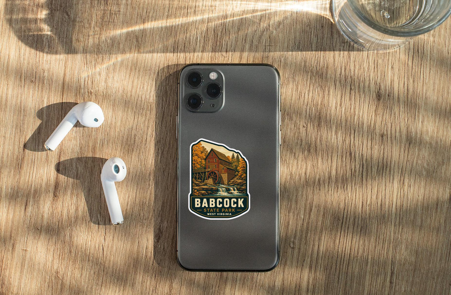 a smartphone with a sticker on it, placed on a wooden surface alongside a pair of white earbuds and a glass of water.