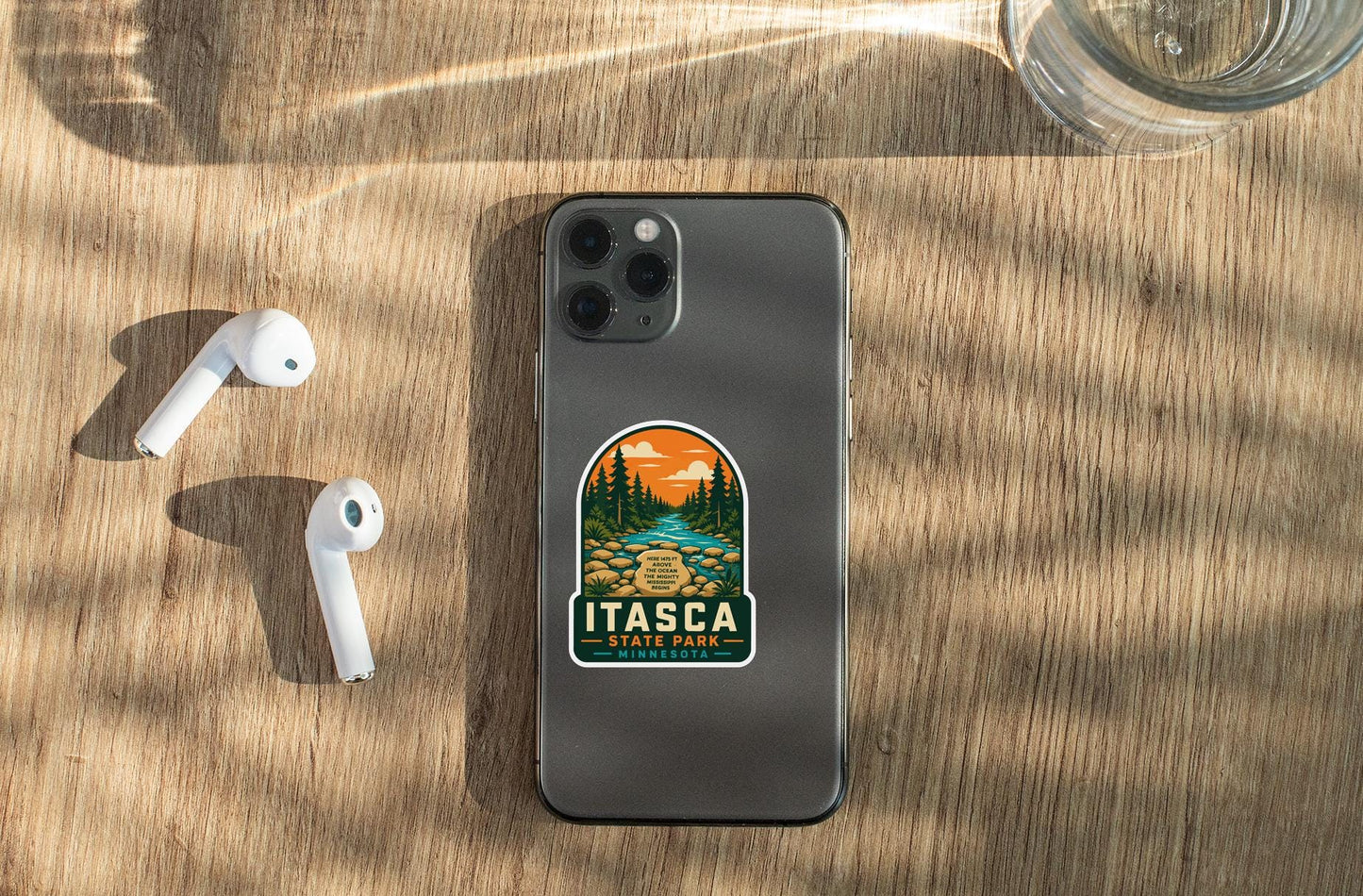 a smartphone with a sticker on the back, placed on a wooden surface alongside a pair of white earbuds and a glass of water.