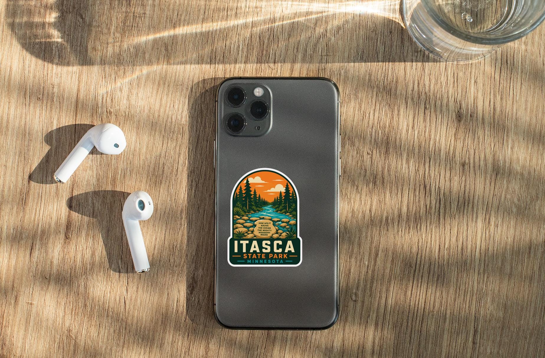 a smartphone with a sticker on the back, placed on a wooden surface alongside a pair of white earbuds and a glass of water.