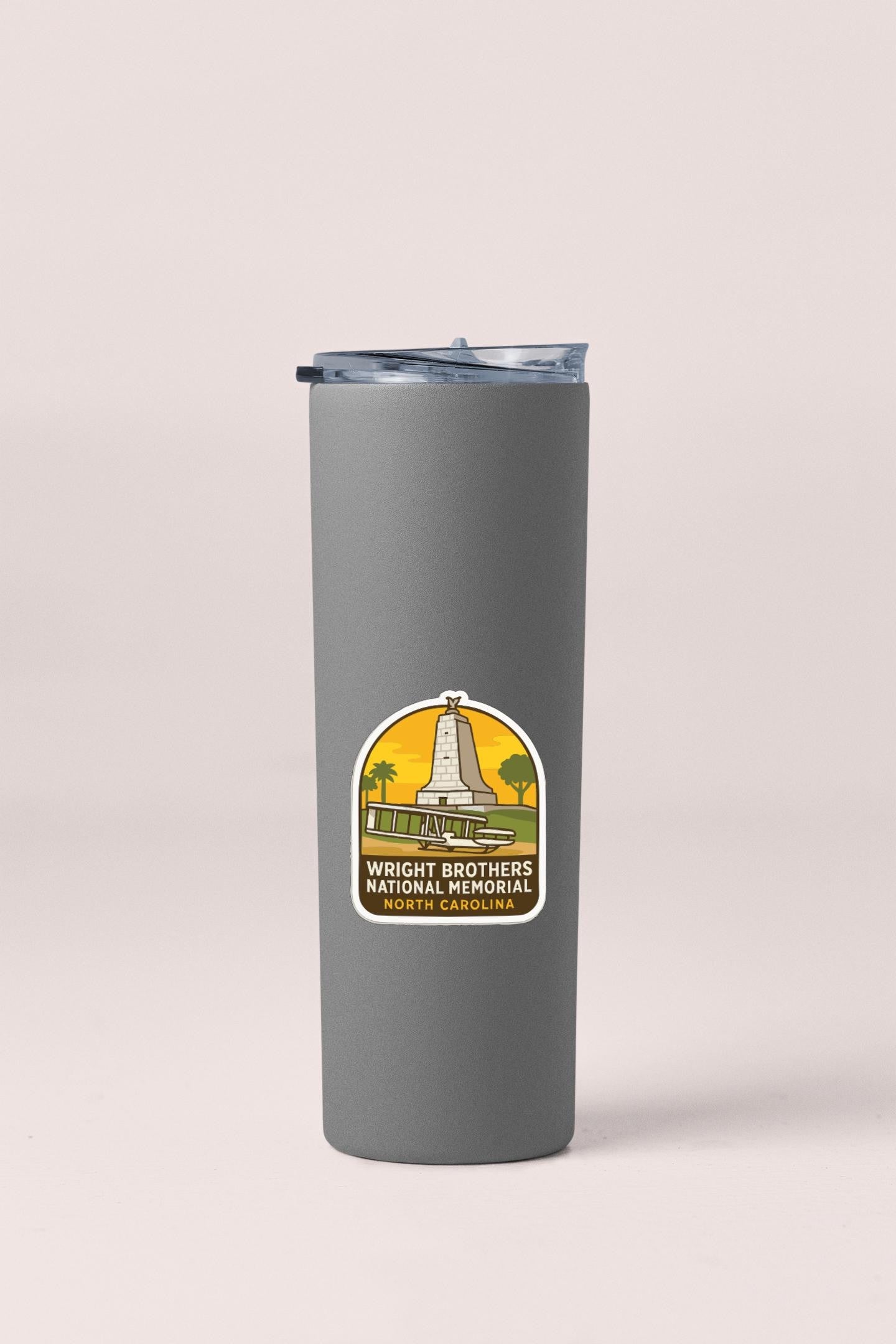 A gray insulated tumbler with a sticker on it, featuring an illustration of a monument and text that says "Wright Brothers National Memorial North Carolina".