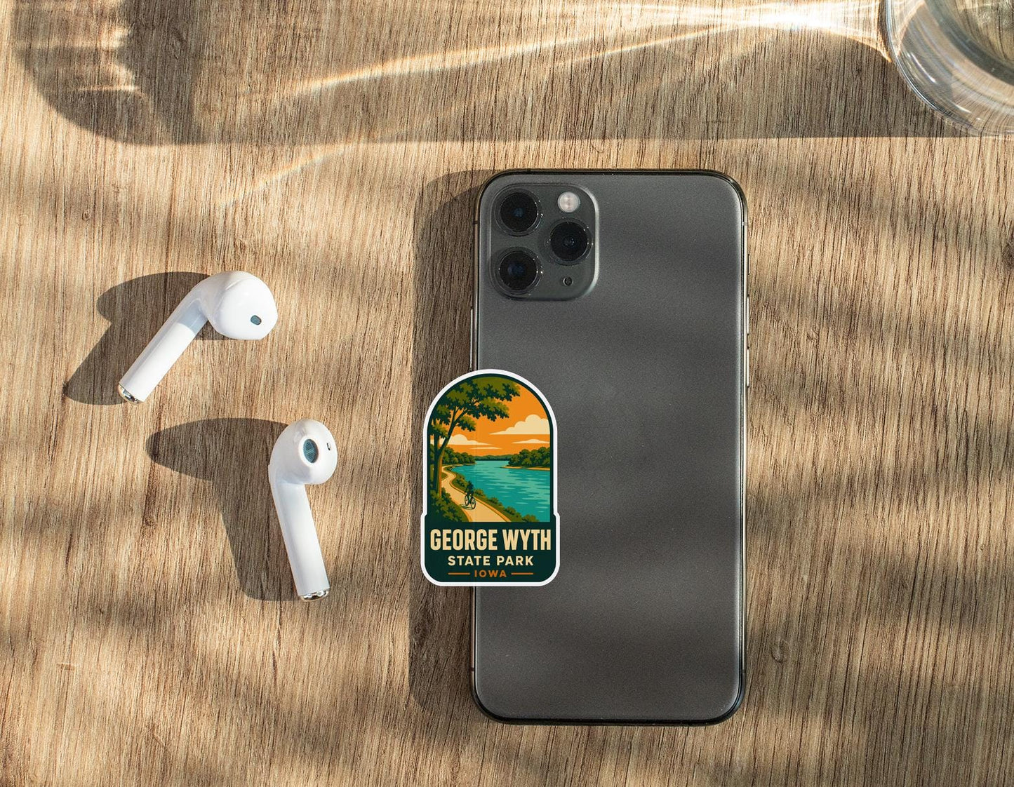 a smartphone with a sticker on it, along with a pair of white earbuds and a glass of water on a wooden surface.