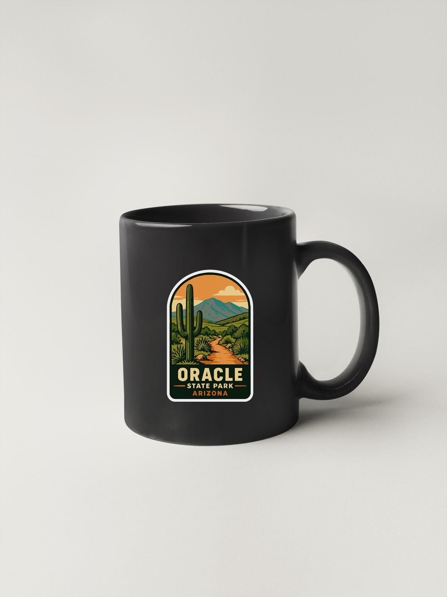A black coffee mug with a graphic design depicting a desert landscape with a cactus and mountains, along with the text "ORACLE STATE PARK ARIZONA".