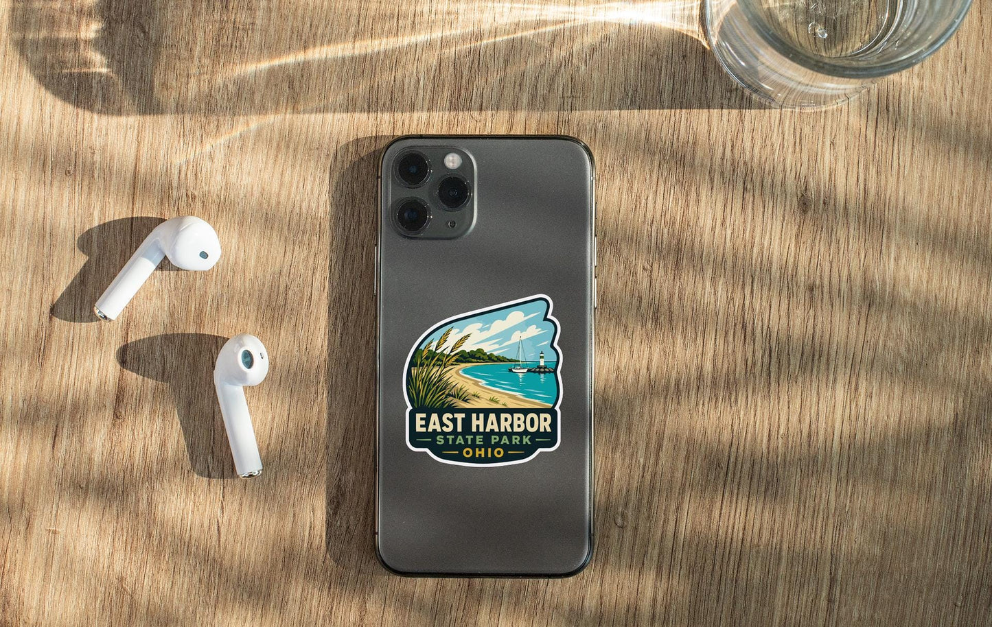 a smartphone with a sticker on the back, placed on a wooden surface alongside a pair of white earbuds and a glass of water.