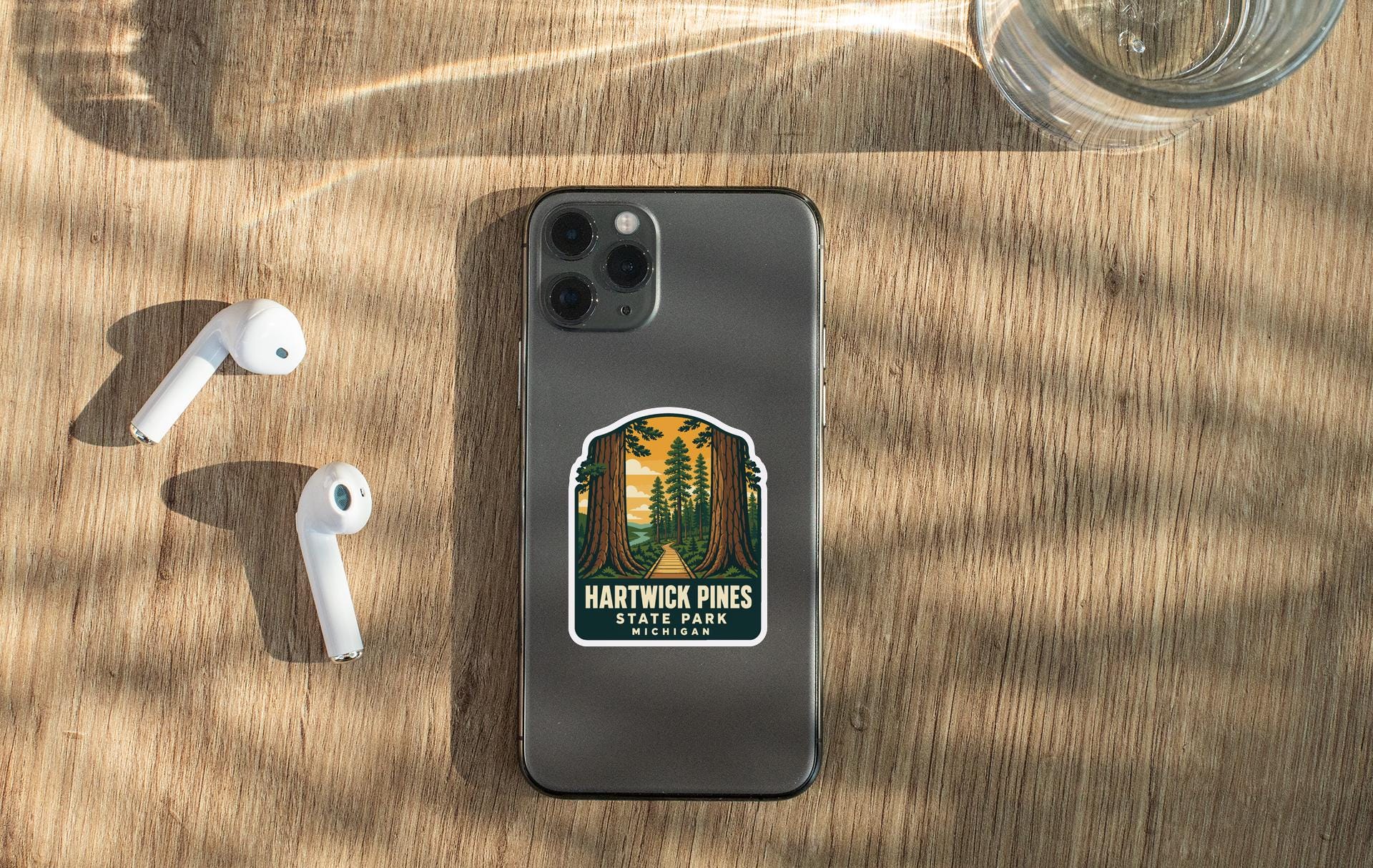a smartphone with a sticker on the back, a pair of white earbuds, and a glass of water on a wooden surface.